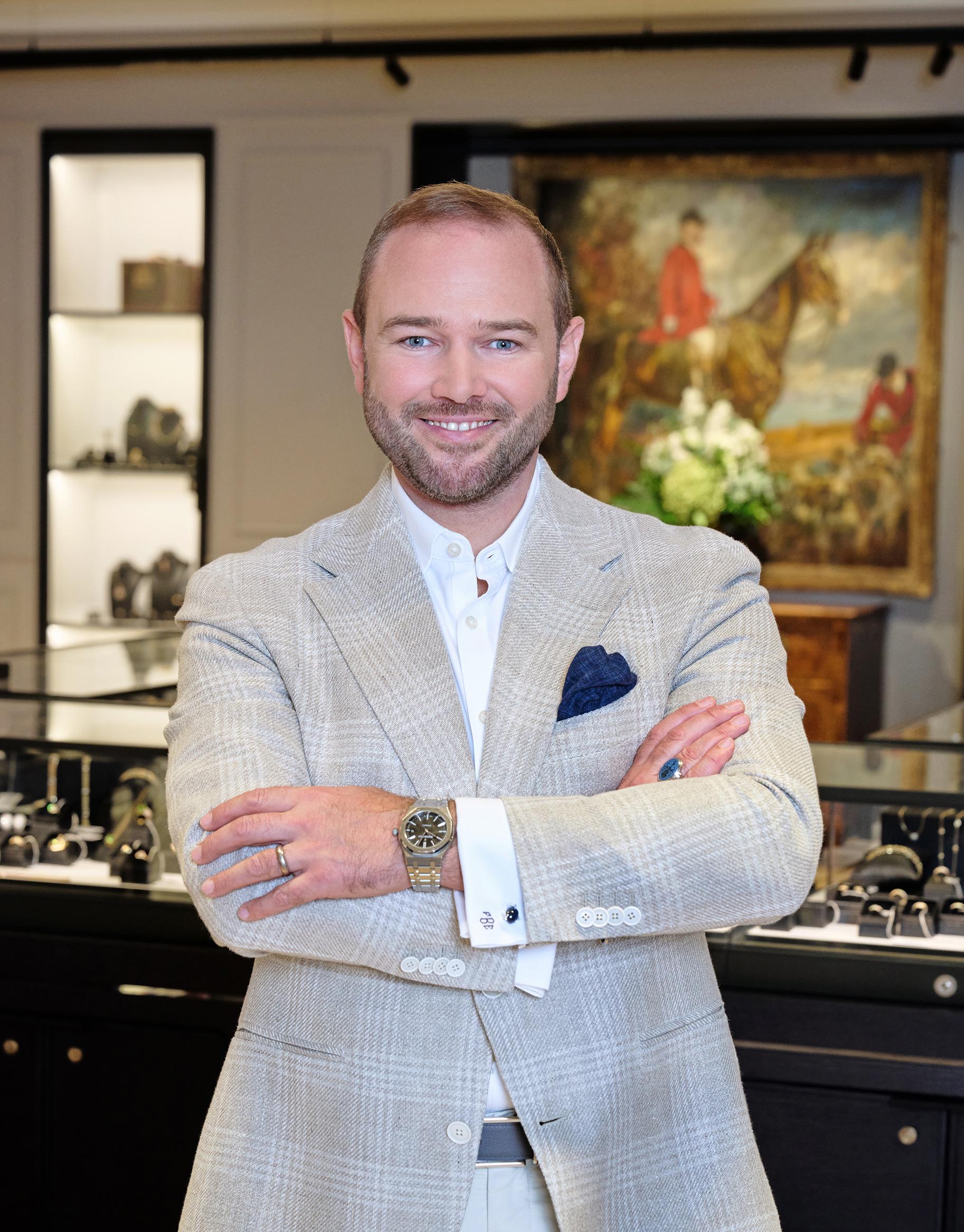 Portrait of Ferdinand Eppli standing with his arms folded inside an auction house showroom displaying jewellery and watches