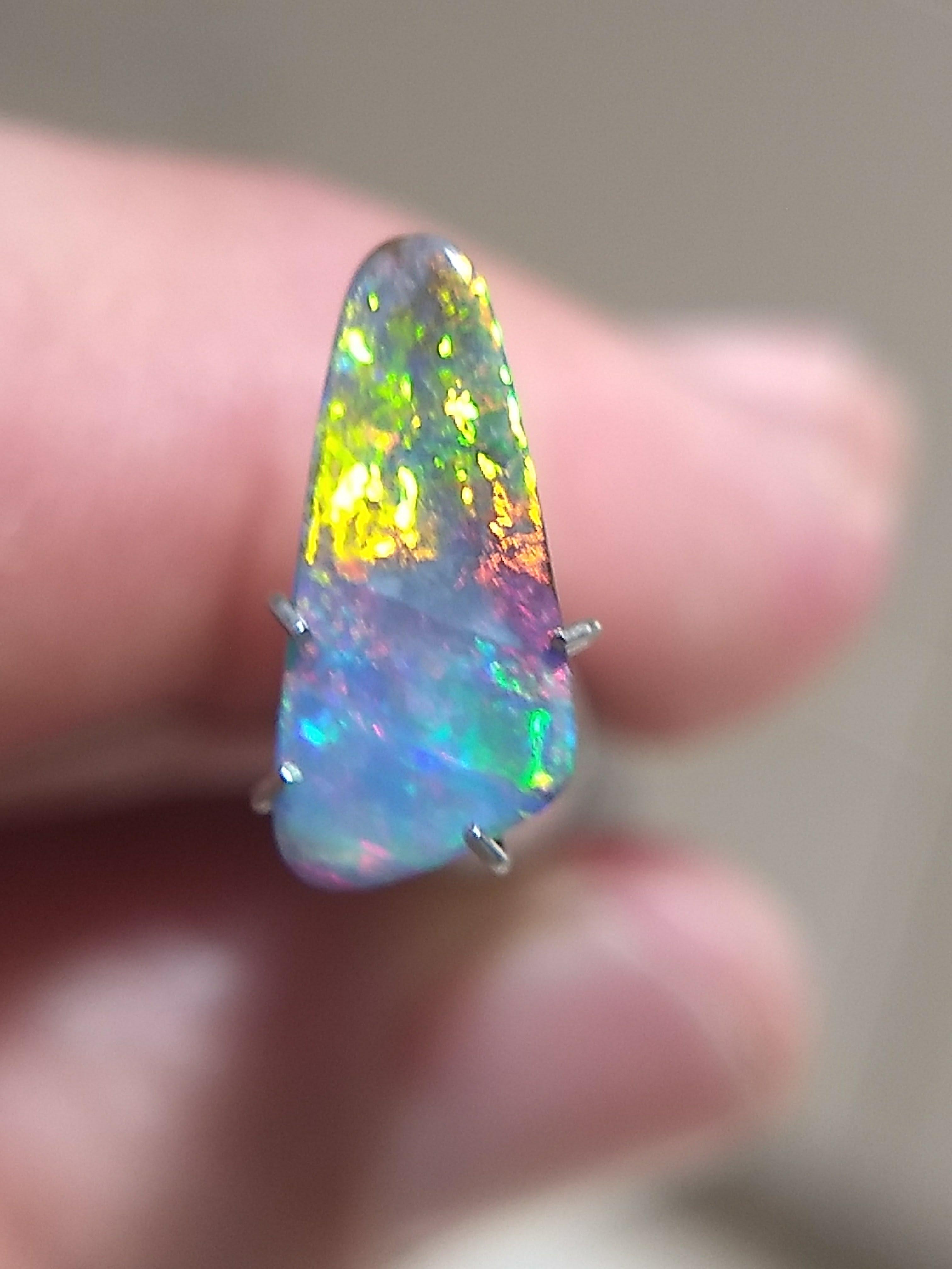 Close-up of an elongated freeform opal held between fingers, displaying vivid green, yellow, blue and pink play-of-colour