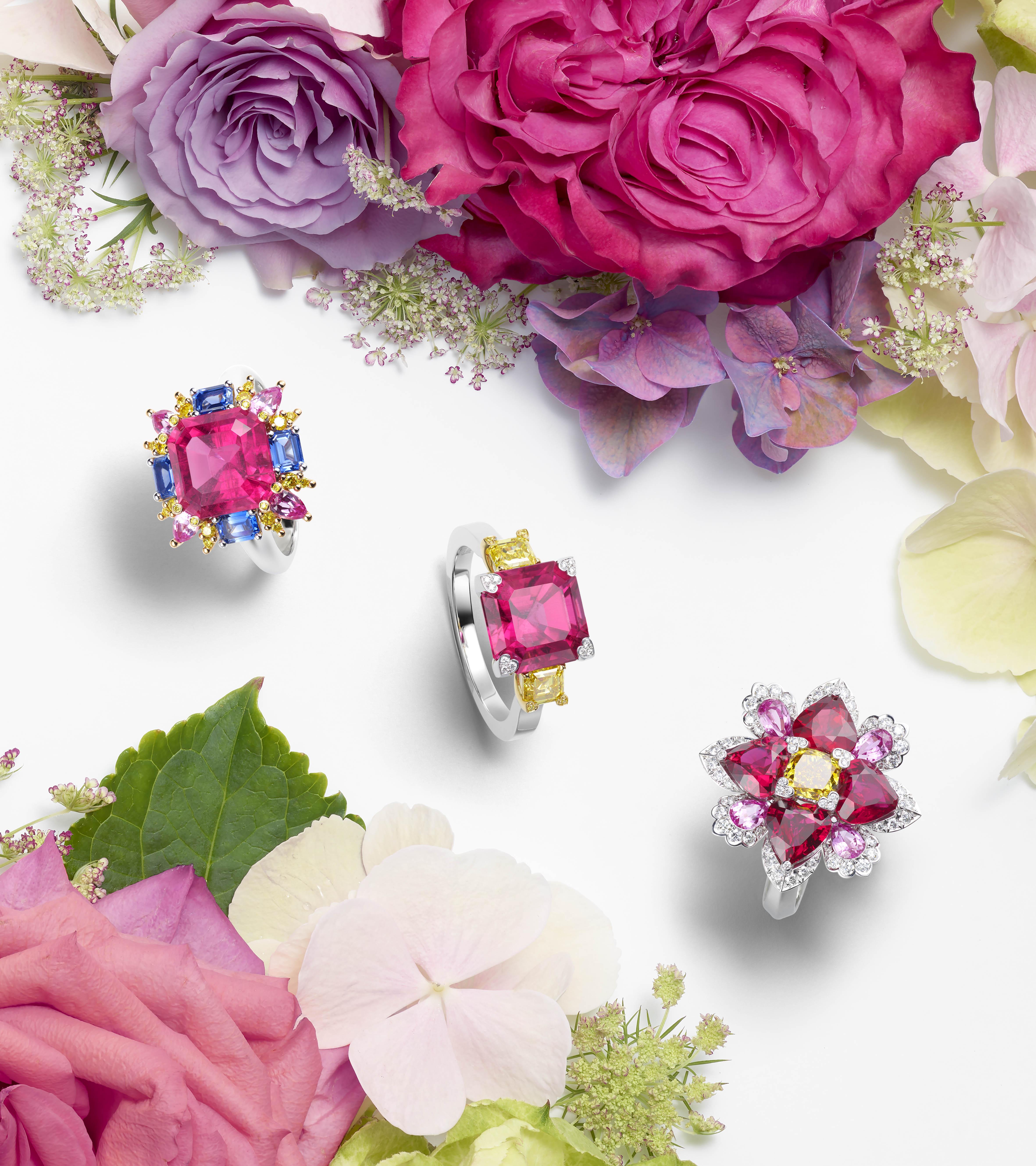 Three gemstone rings with pink and blue stones and diamonds arranged on a background of colourful flowers