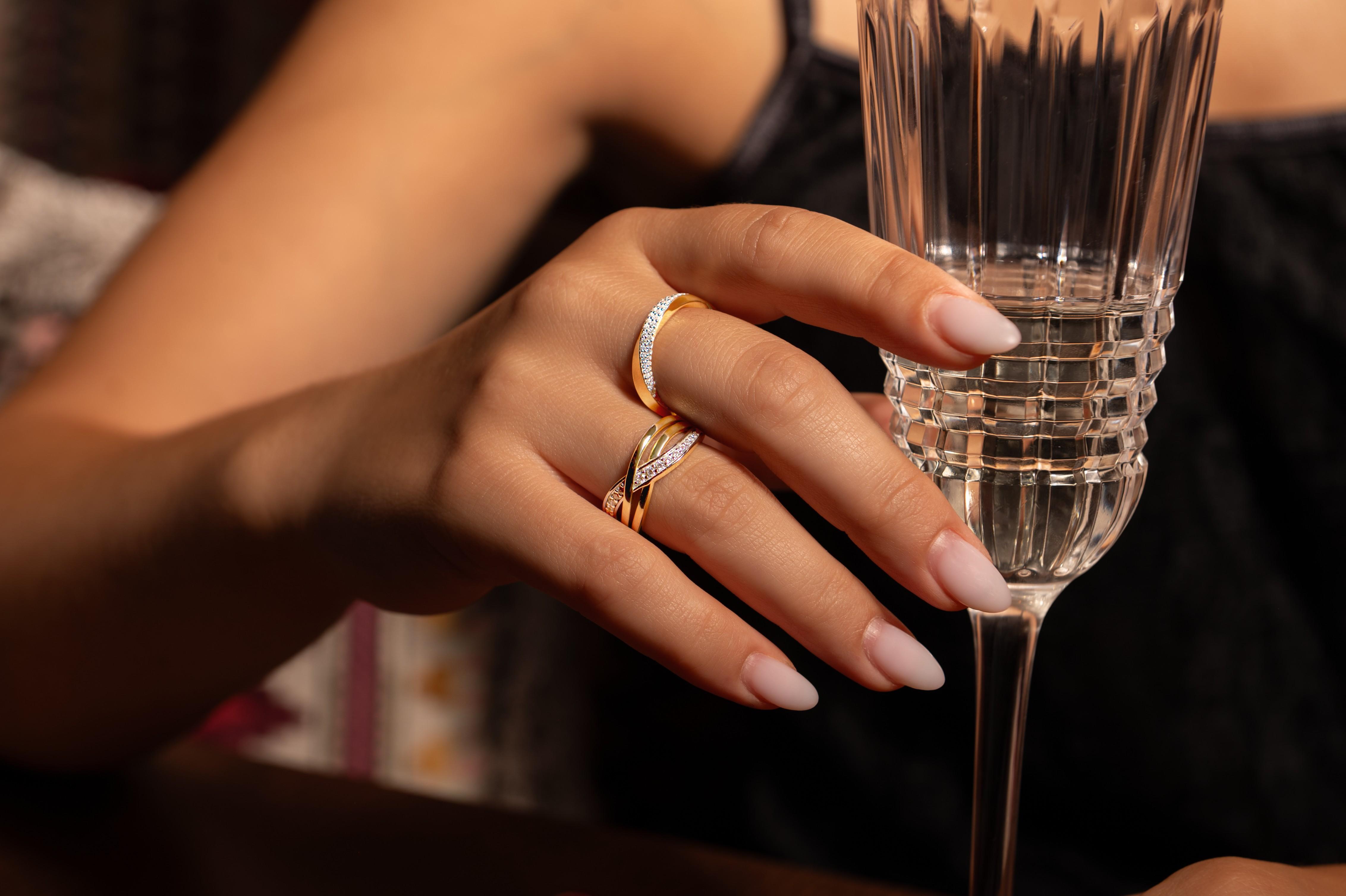 Close-up of a hand wearing two Gisser gold-plated rings with diamond detailing beside a champagne flute.