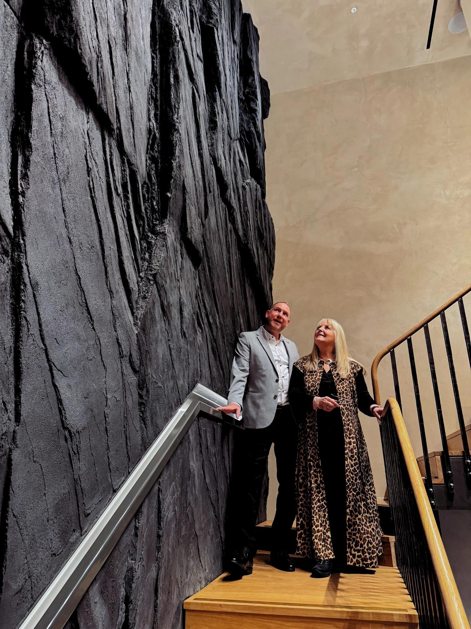 Sheila Fleet and a project collaborator stand on the wooden staircase beside the gallery’s towering dark rock-textured wall.