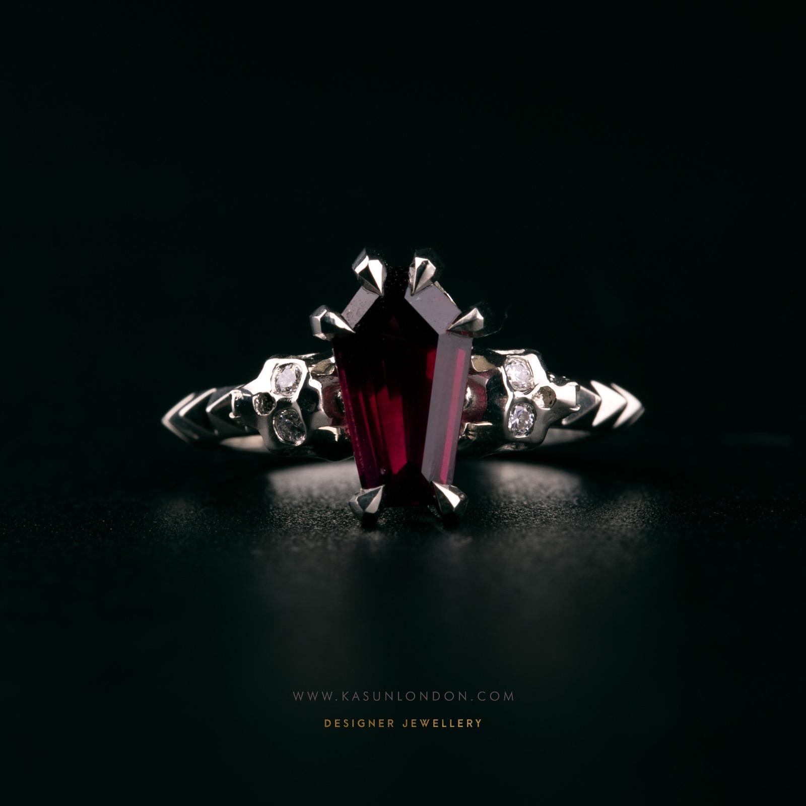 Front view of a bespoke white gold engagement ring with a deep red coffin-cut garnet, sculpted skulls set with diamonds and pointed geometric shoulders.