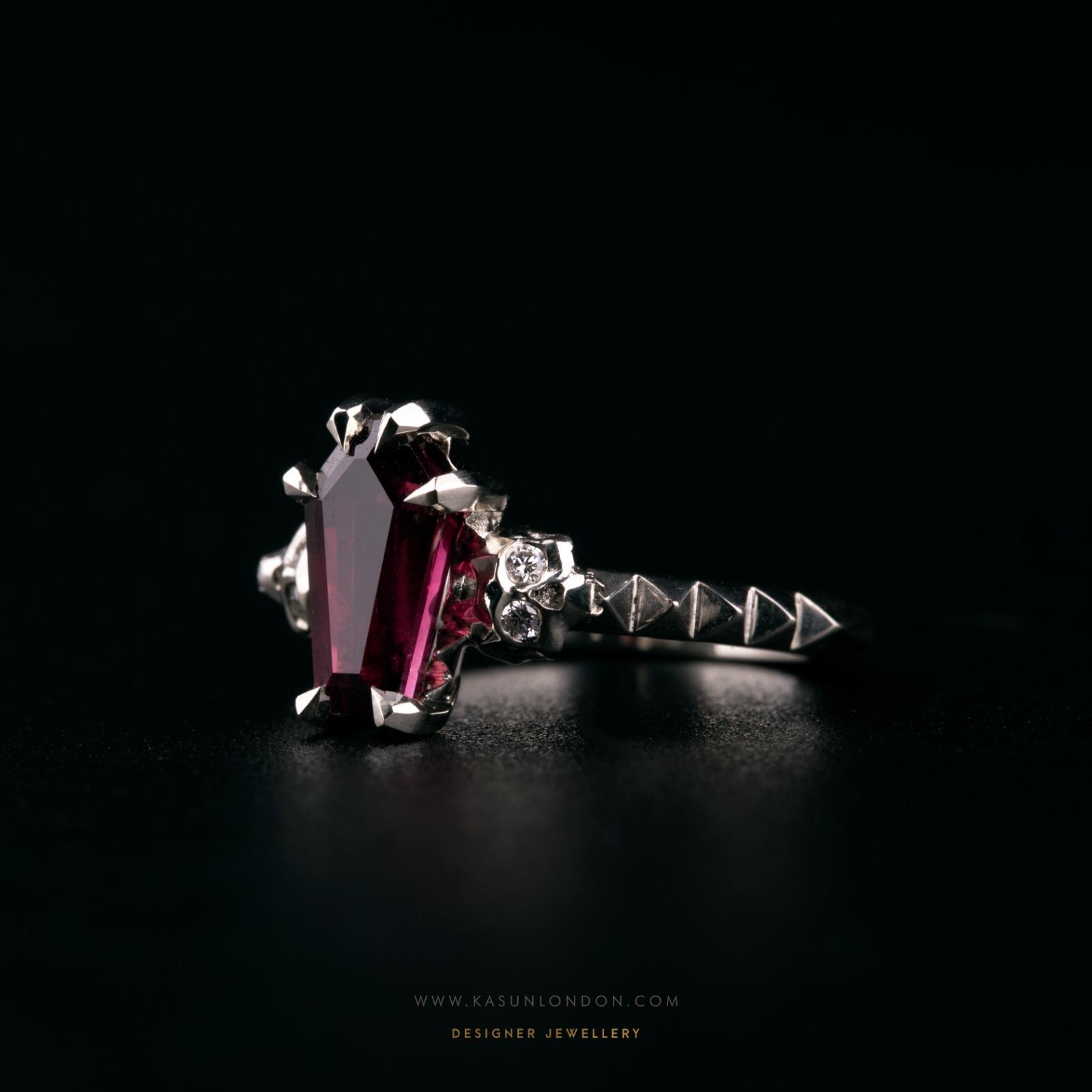 Side view of a bespoke white gold engagement ring with a deep red coffin-cut garnet, a diamond-set skull and pointed geometric details along the band
