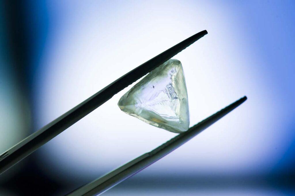 Triangular transparent rough diamond held between black tweezers against a blue-and-white background.