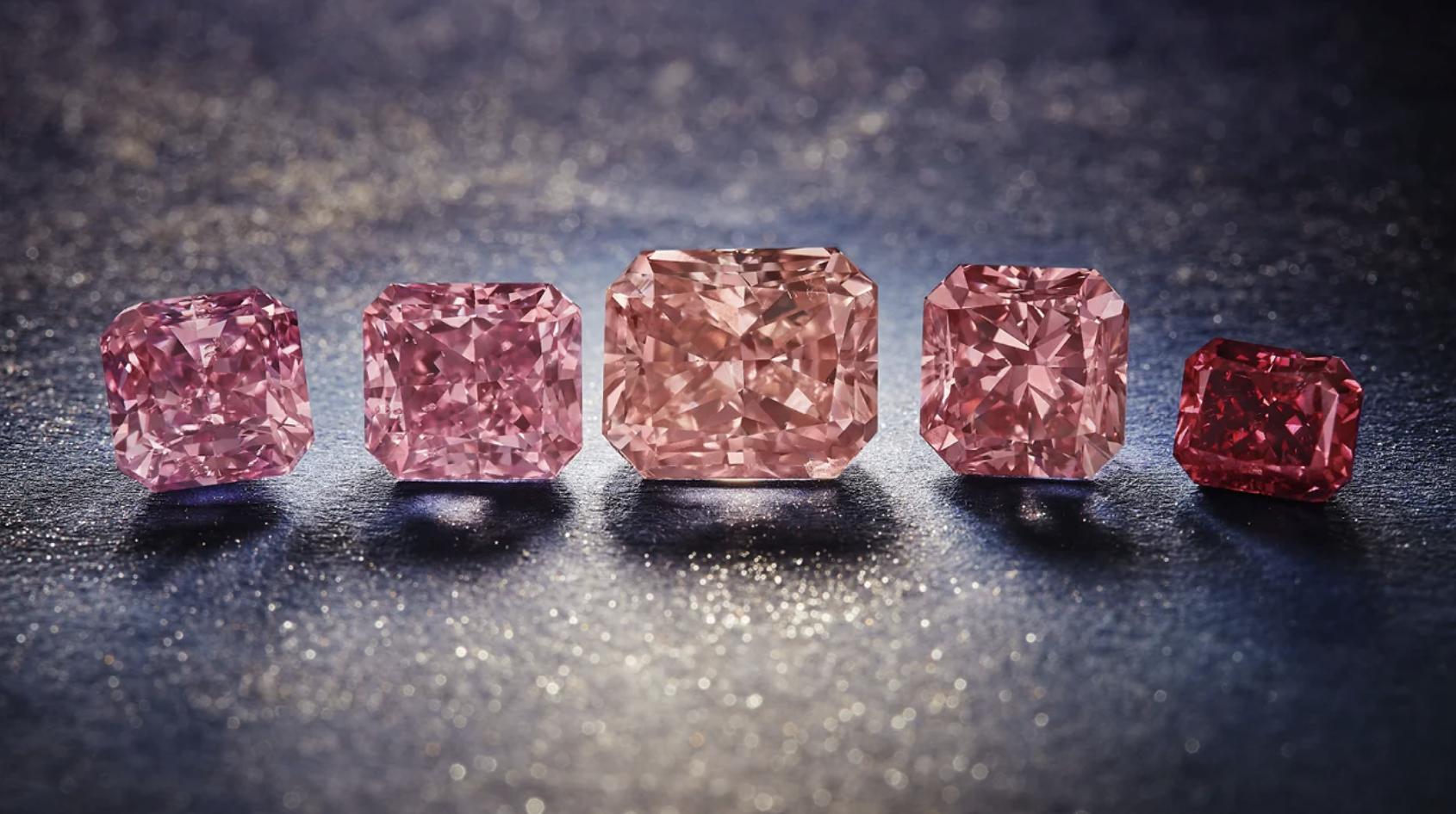 Five rectangular-cut Argyle diamonds in shades ranging from pale pink to deep red, arranged on a dark reflective surface
