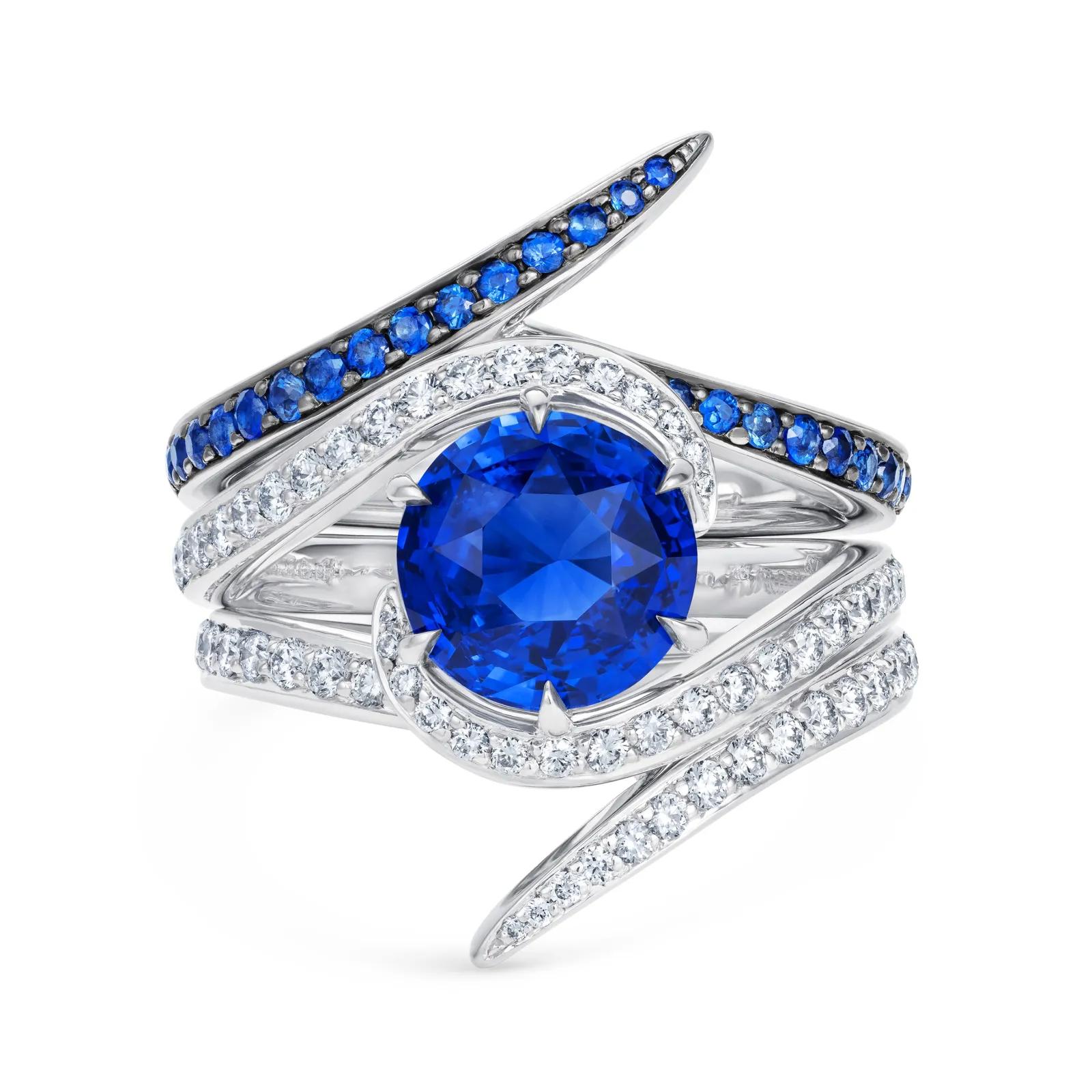 Sculptural white-metal ring with a large round blue gemstone surrounded by curved rows of diamonds and smaller blue stones.
