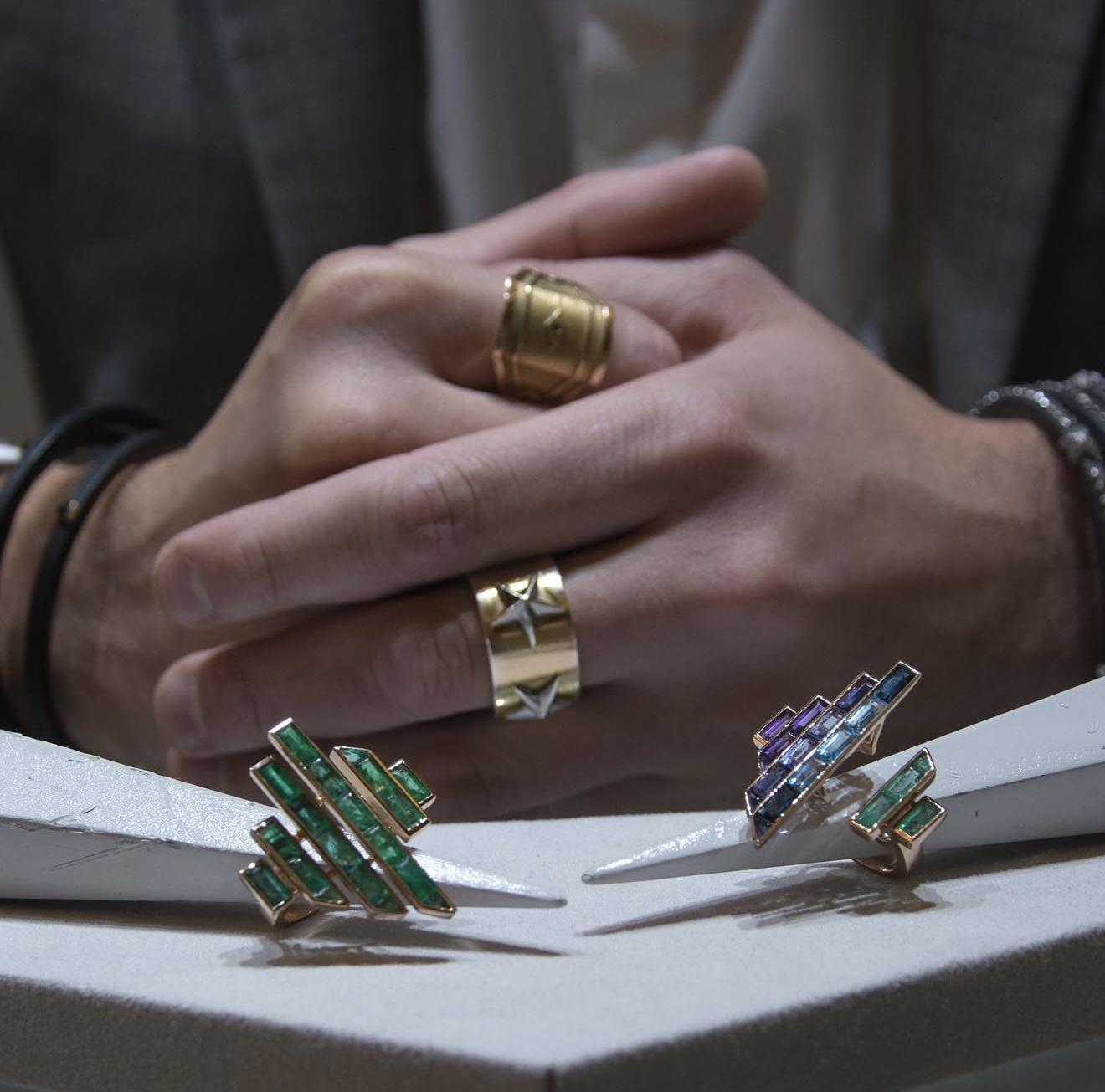 Emerald, blue and violet gemstone rings by Tomasz Donocik displayed in front of hands wearing wide sculptural gold rings