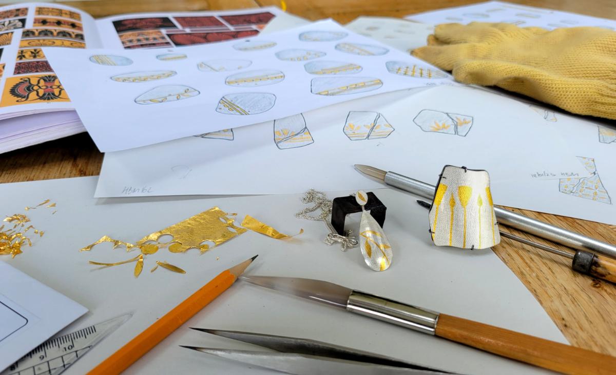 Keum-Boo jewellery samples decorated with gold beside design sketches, sheets of gold foil and jewellery-making tools on a workbench
