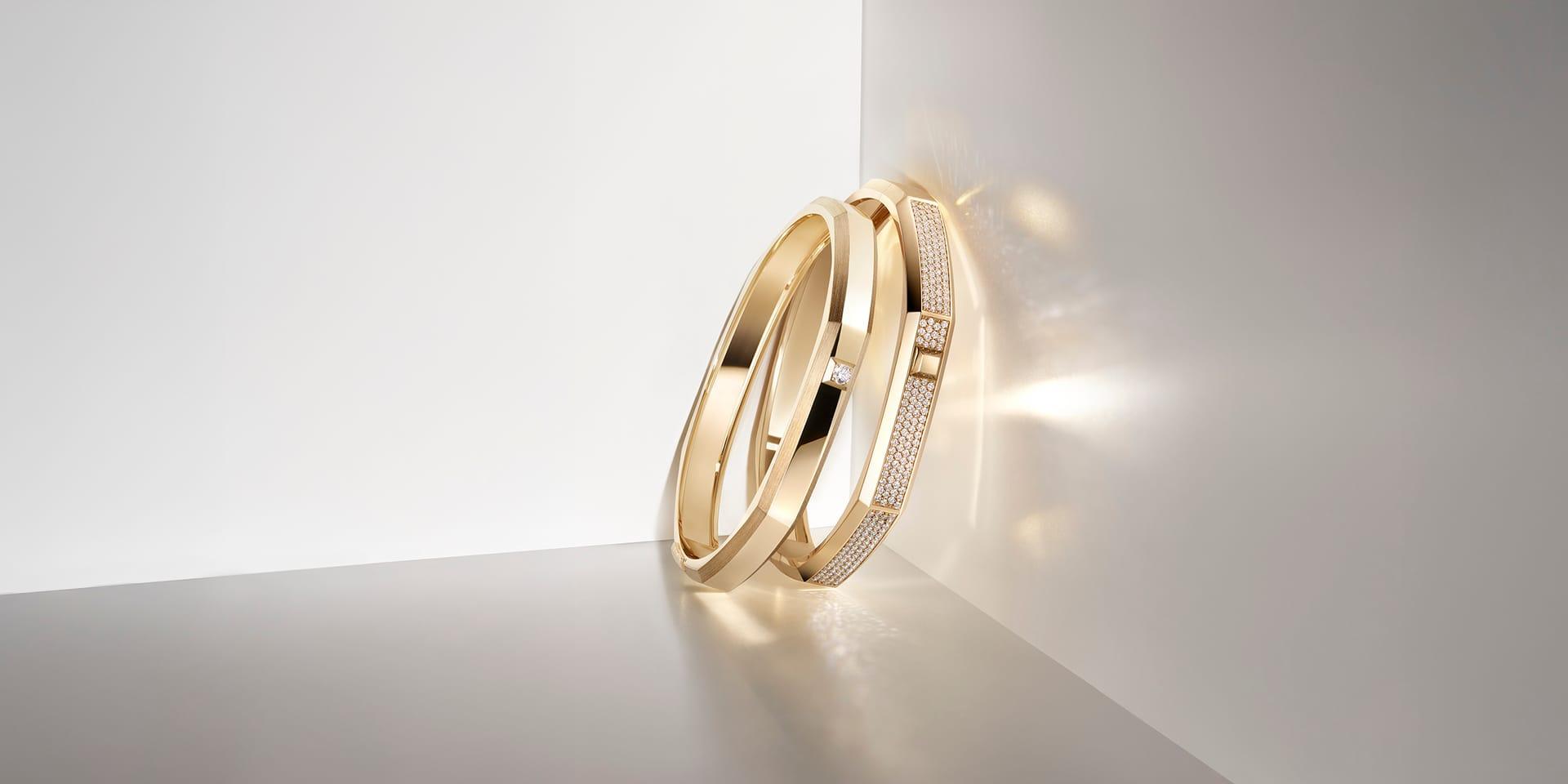Two yellow gold Messika Moderniste bangles leaning against a reflective surface, featuring polished geometric lines and pavé diamond sections illuminated by soft light.