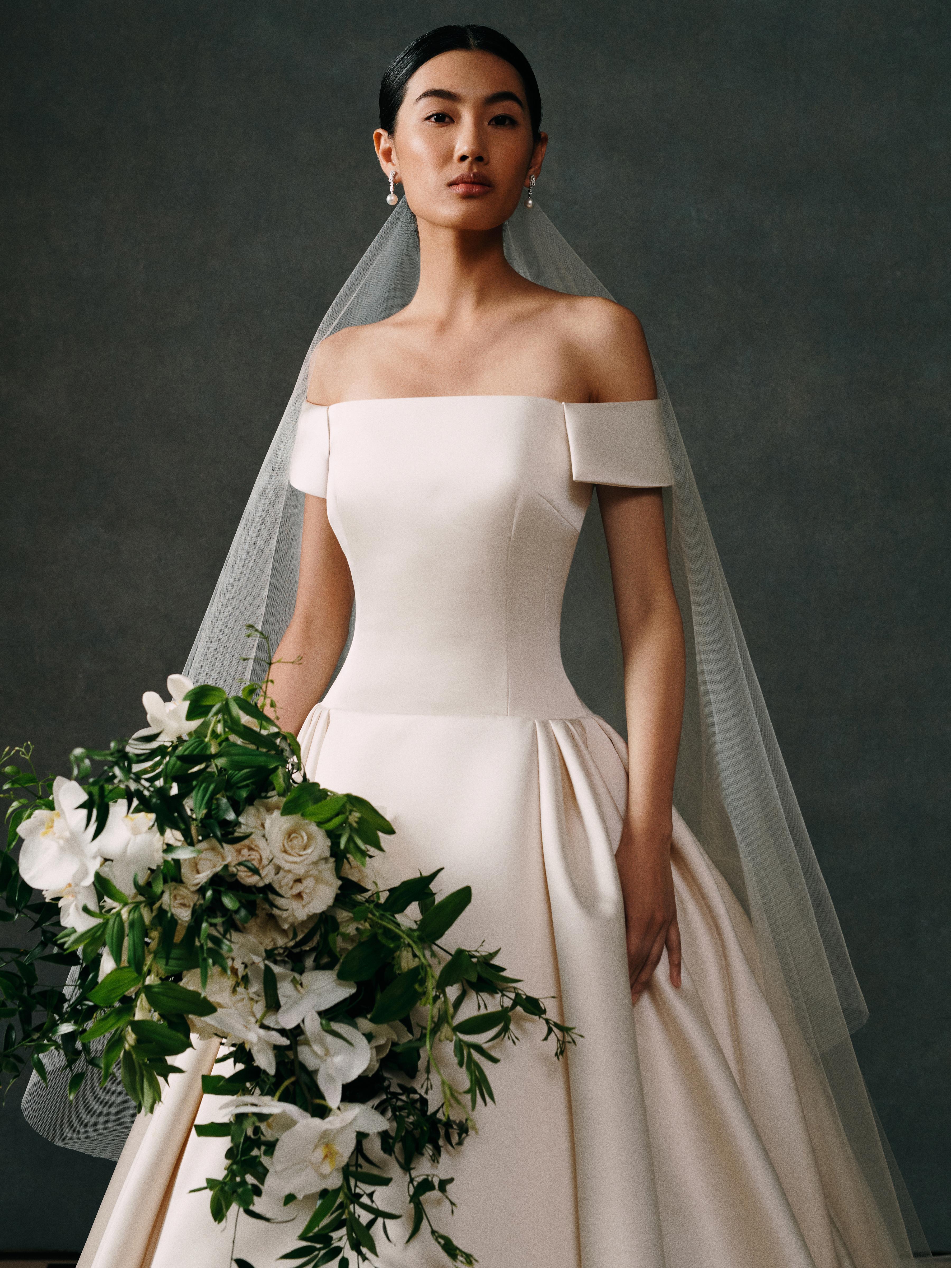Model wearing an off-the-shoulder bridal gown and veil, styled with pearl drop earrings from the Yoko London x Emilia Wickstead bridal collection and holding a bouquet of white flowers and greenery