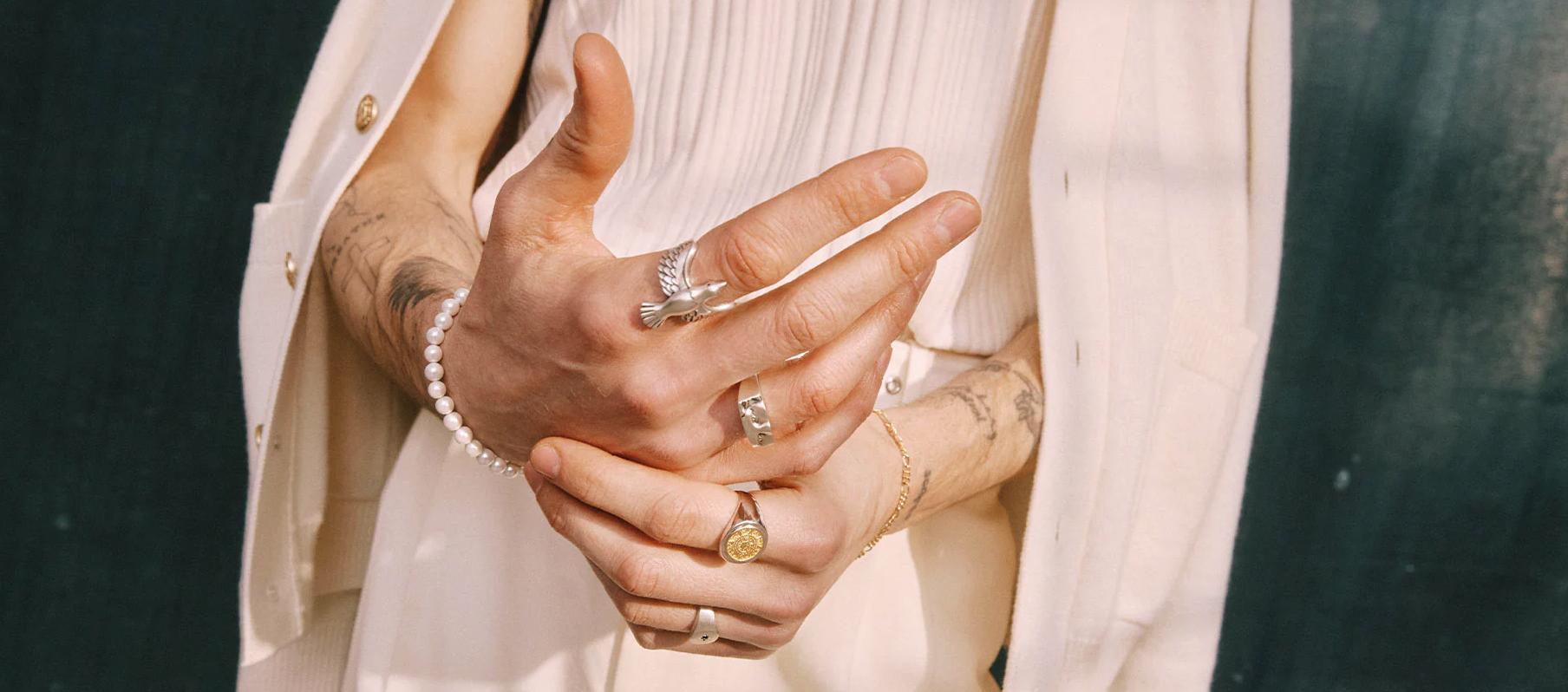Close-up of hands wearing multiple Serge Denimes rings and bracelets, including a signet ring and beaded bracelet, styled with neutral clothing.