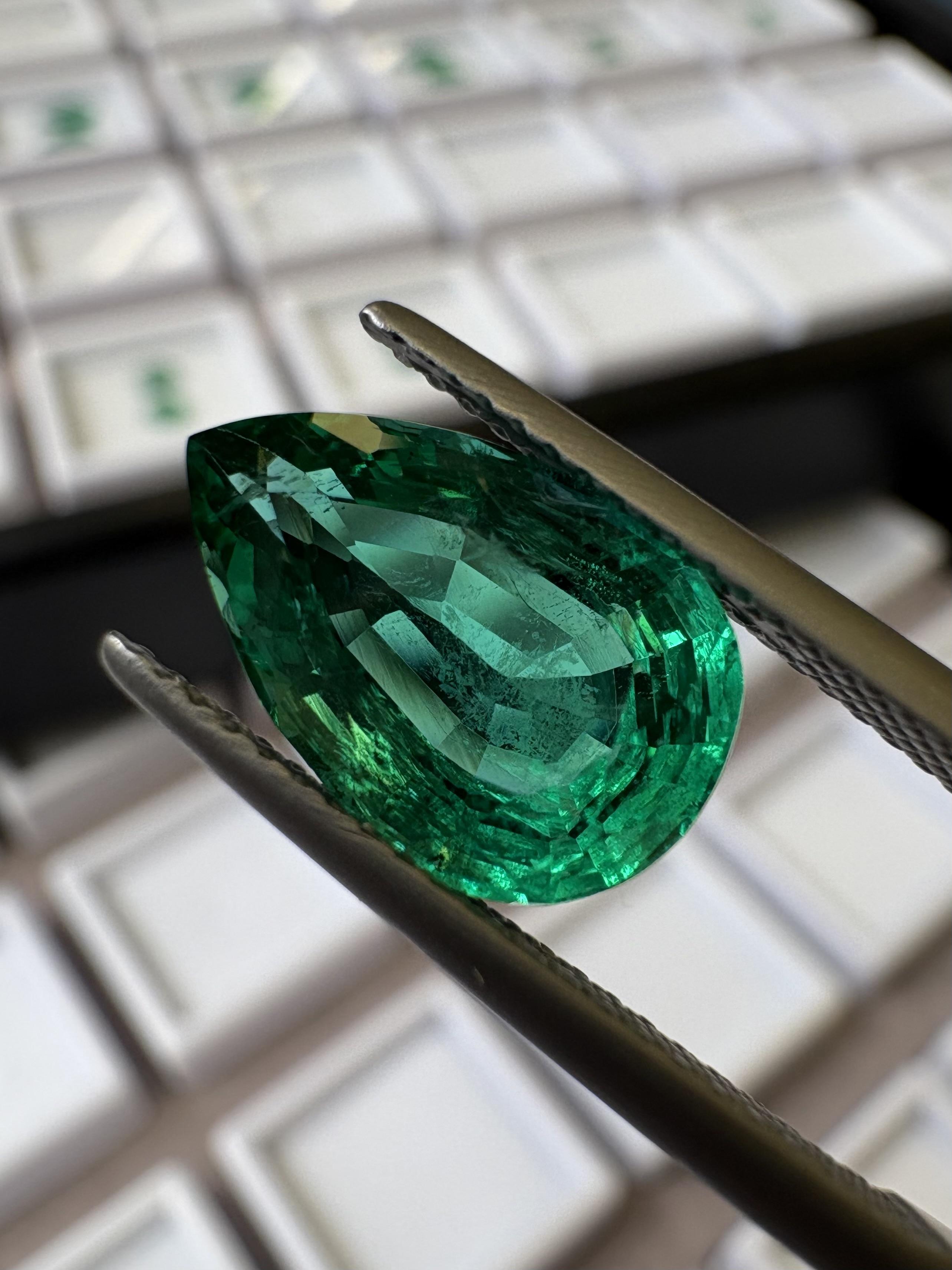 Close-up of a pear-cut emerald held in tweezers with gemstone parcels blurred in the background.