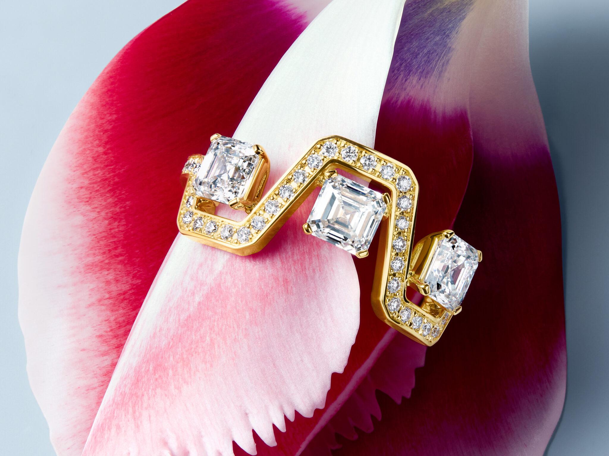 Pair of yellow gold and diamond Skyline earrings featuring angular geometric forms and emerald-cut diamonds, displayed against vibrant pink and white flower petals.