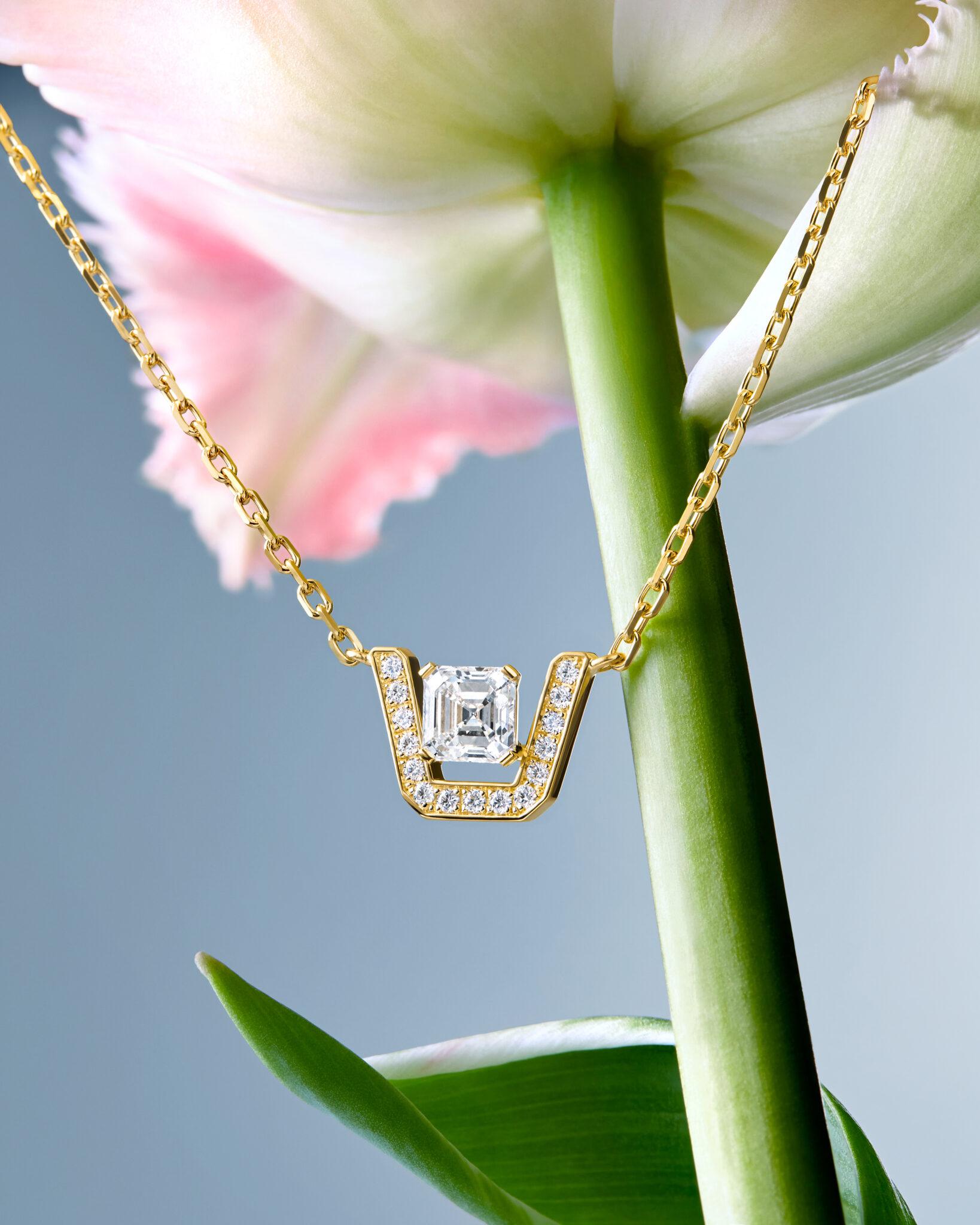 Yellow gold Skyline necklace featuring an asscher-cut diamond surrounded by pav&eacute;-set diamonds, suspended against the stem and petals of a pink and white flower.