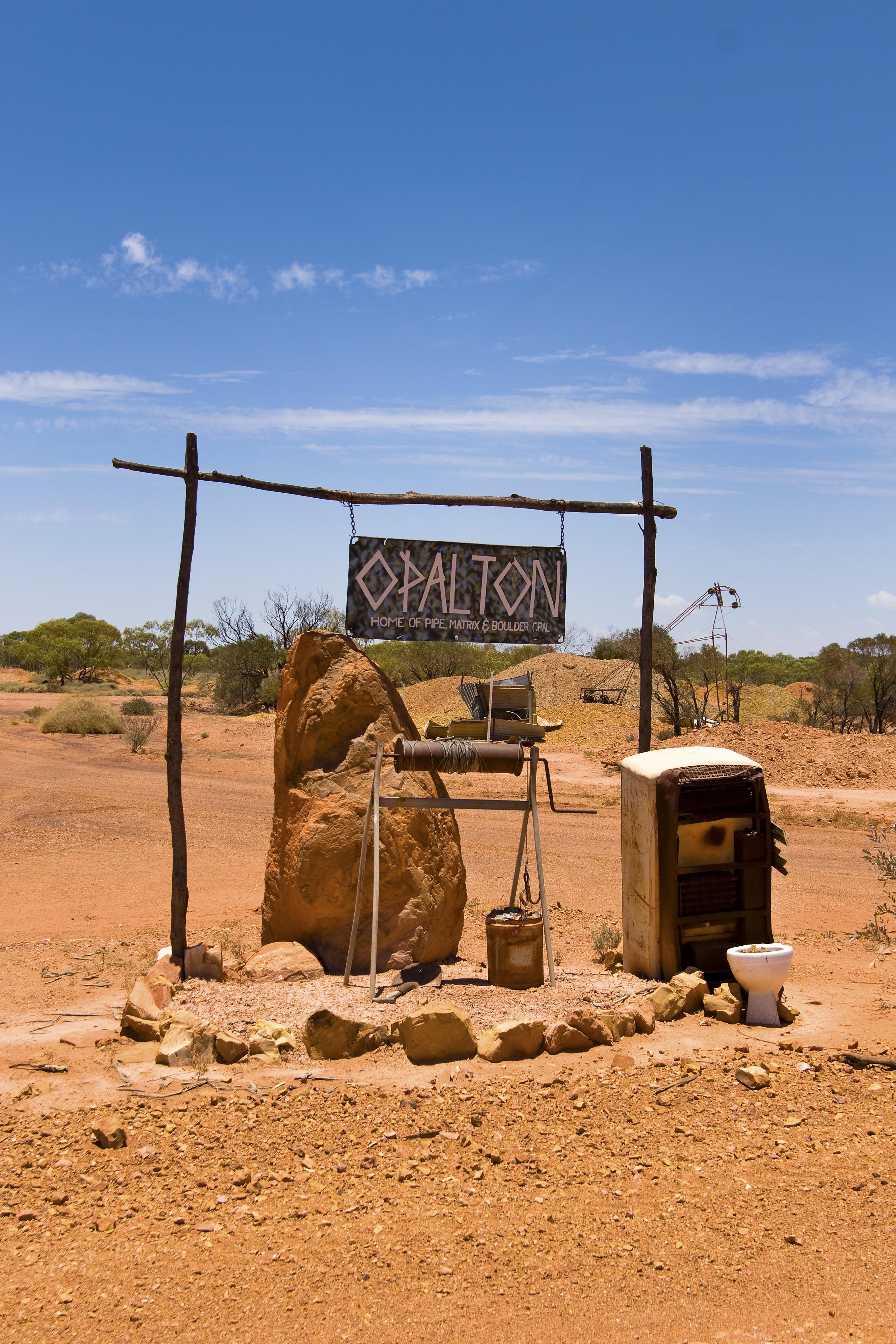 A rustic Opalton sign, stone well and mailbox standing in the dry red landscape of western Queensland beneath a blue sky