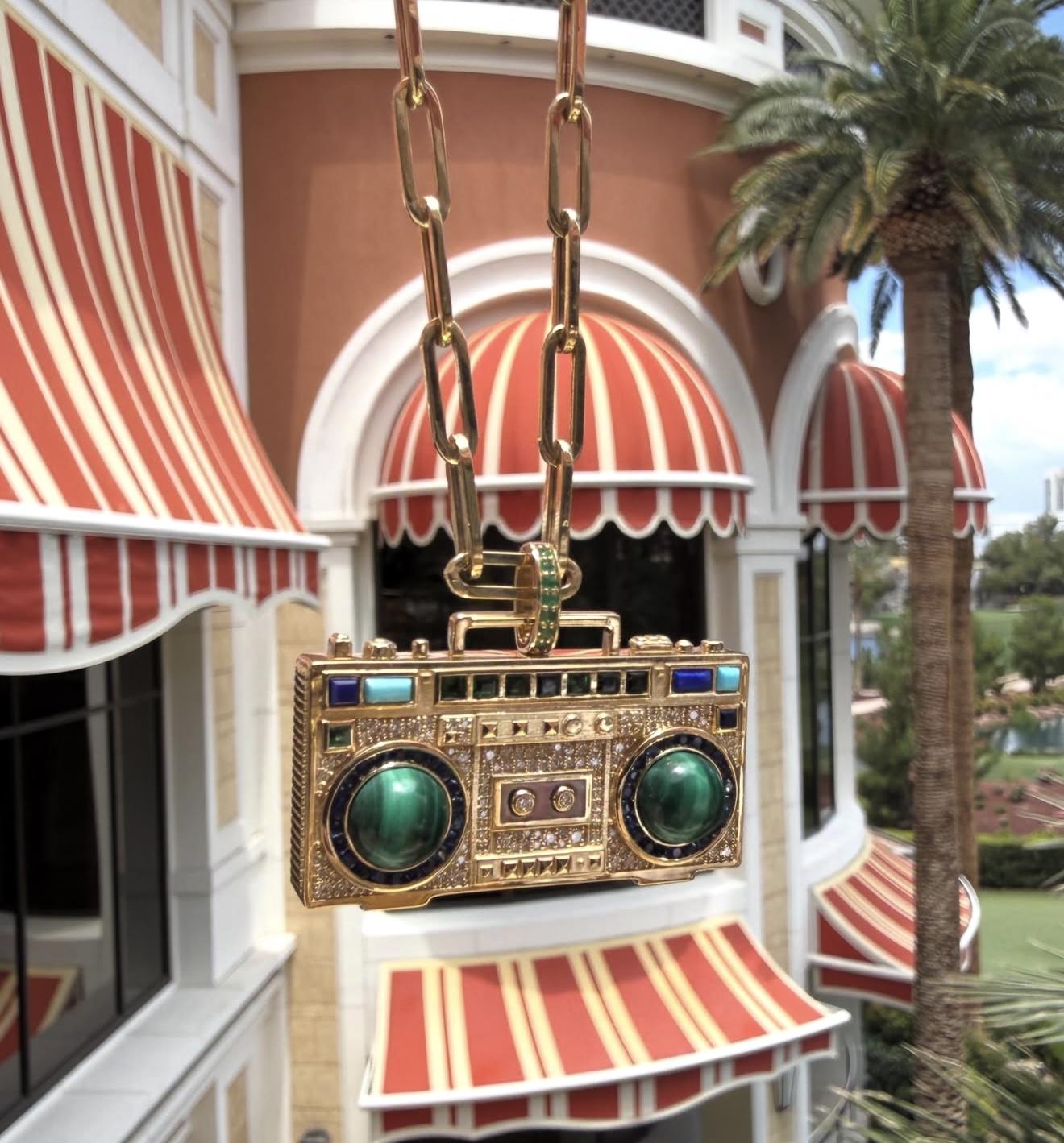 Close-up of a NeverNoT Turn It Up yellow gold boombox pendant set with diamonds, malachite and coloured gemstones displayed outdoors.