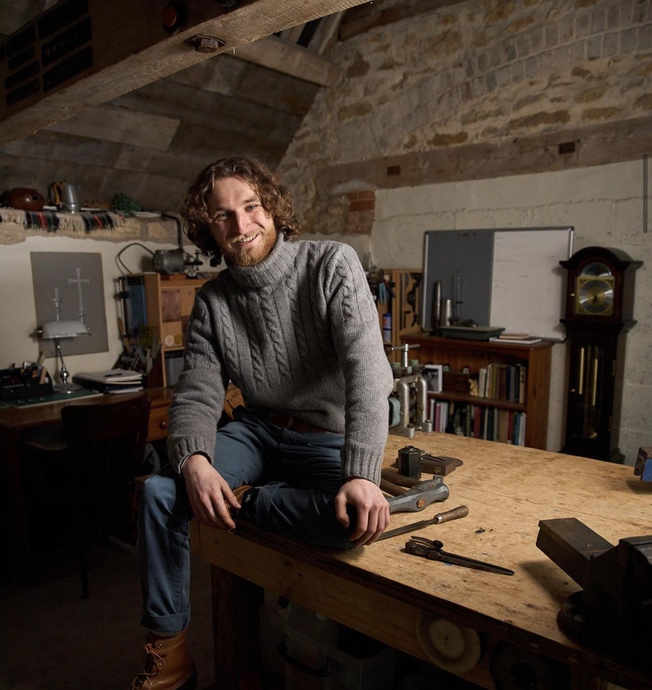 Silversmith Harry Stokes sitting on a wooden workbench surrounded by hand tools in his stone-walled workshop