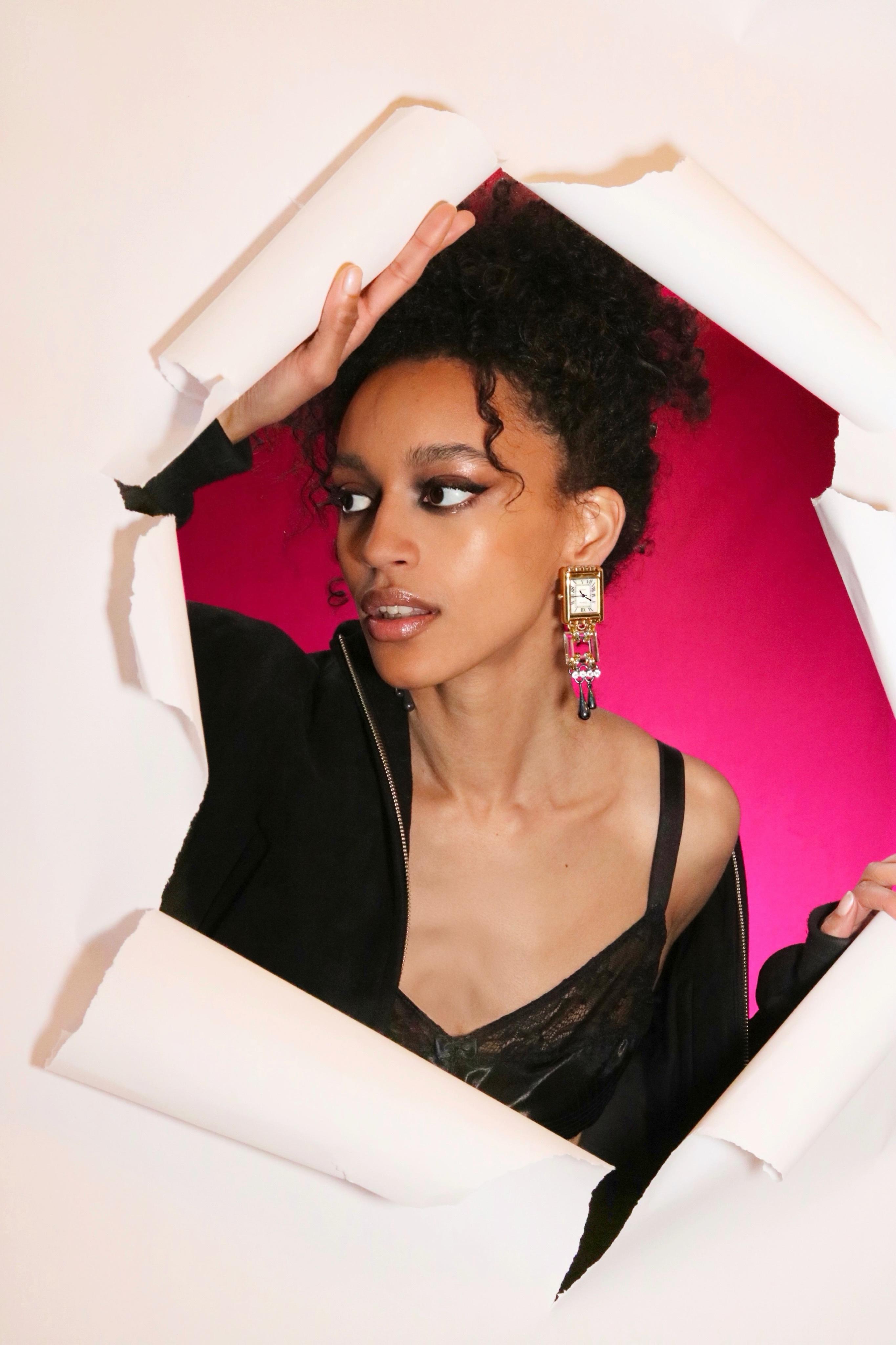 Model wearing statement drop earrings with a vintage watch face, crystals and black detailing against a pink background.