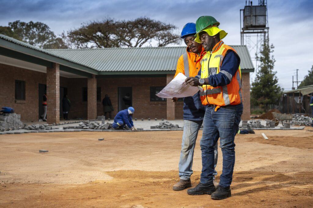 Two construction workers in hard hats and high-visibility clothing reviewing building plans outside a brick development.
