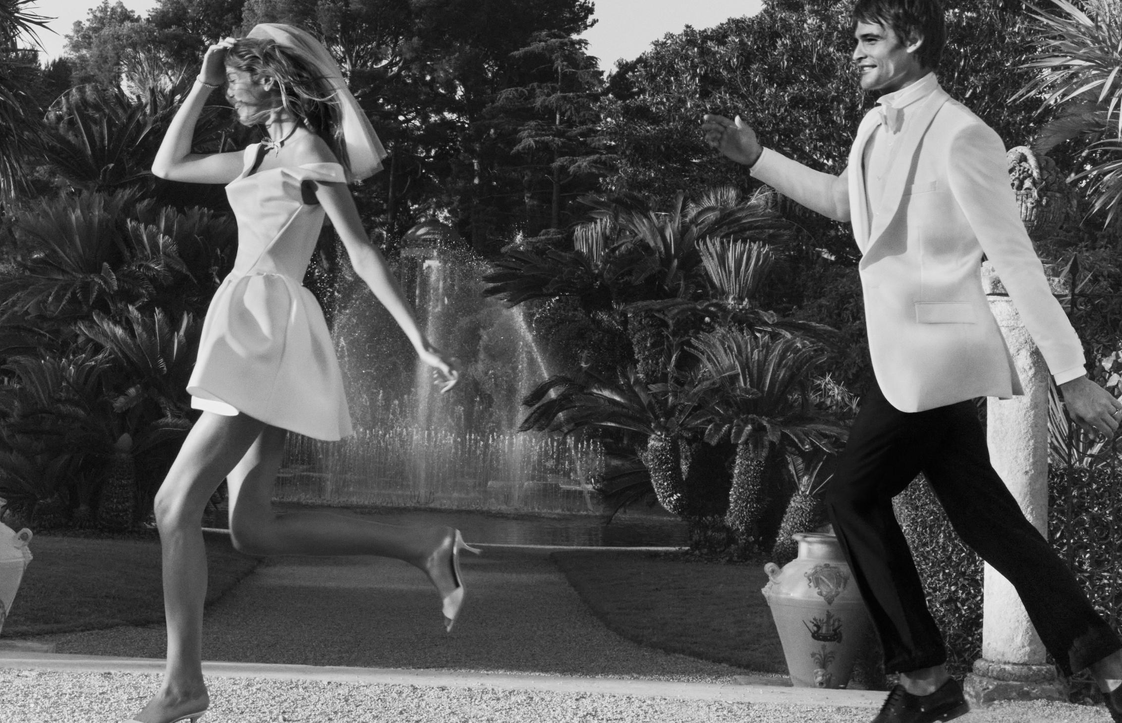 Black-and-white photograph of a bride and groom playfully running through the gardens of a French Riviera villa.