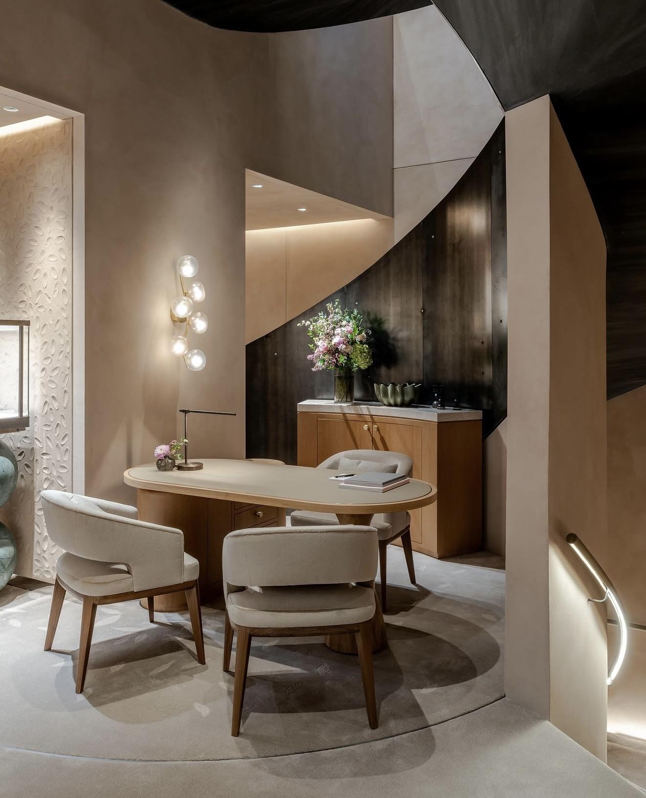 Consultation area inside The Boodles Townhouse with a pale wooden table, upholstered chairs, floral arrangements and a dark sculptural staircase overhead