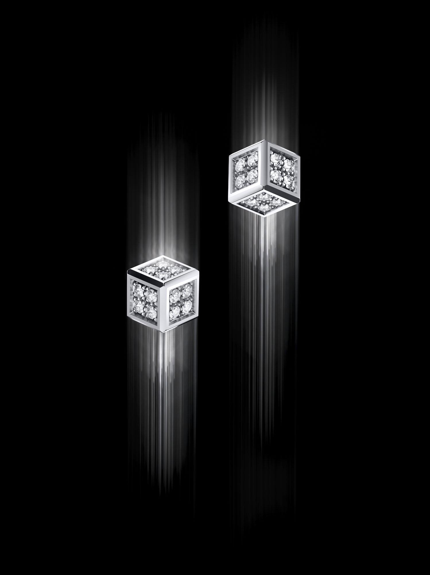 Two diamond-set cube earrings in white metal appear suspended against a black background, with light trails emphasising their geometric form.