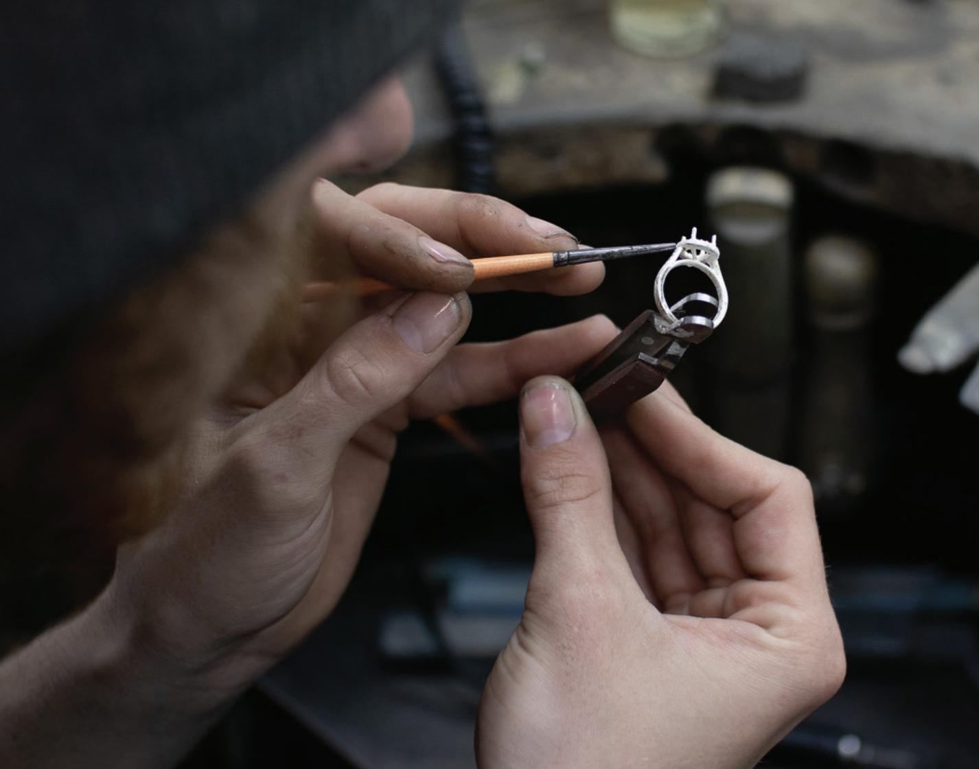 Jewellery craftsperson using a fine brush and pliers to work on an unfinished ring setting at a workshop bench.