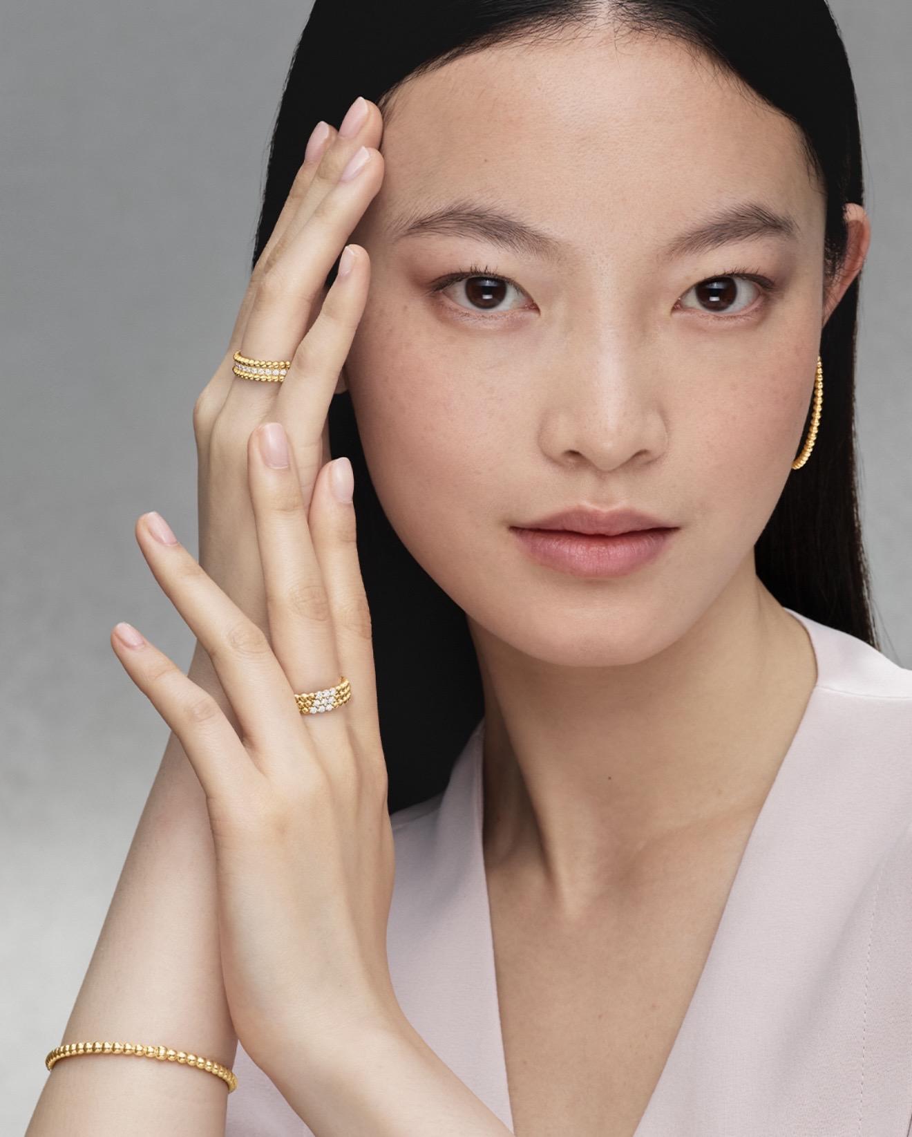 Model wearing Van Cleef & Arpels Perlée yellow gold jewellery including diamond-set rings, hoop earrings and a beaded gold bracelet.