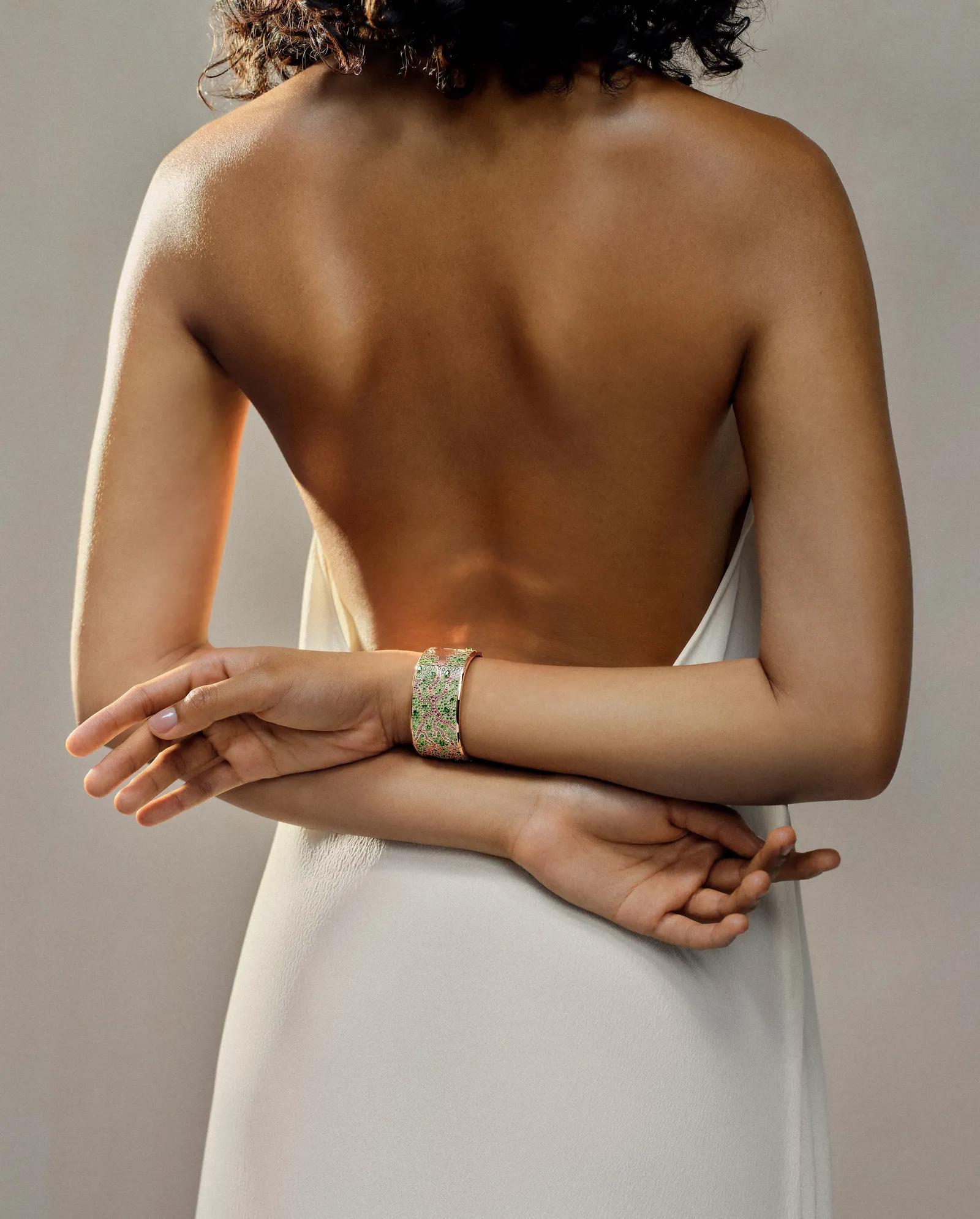 Model in a backless white dress wearing a wide pink-gold Audemars Piguet cuff decorated with an intricate pattern of green, pink and white gemstones