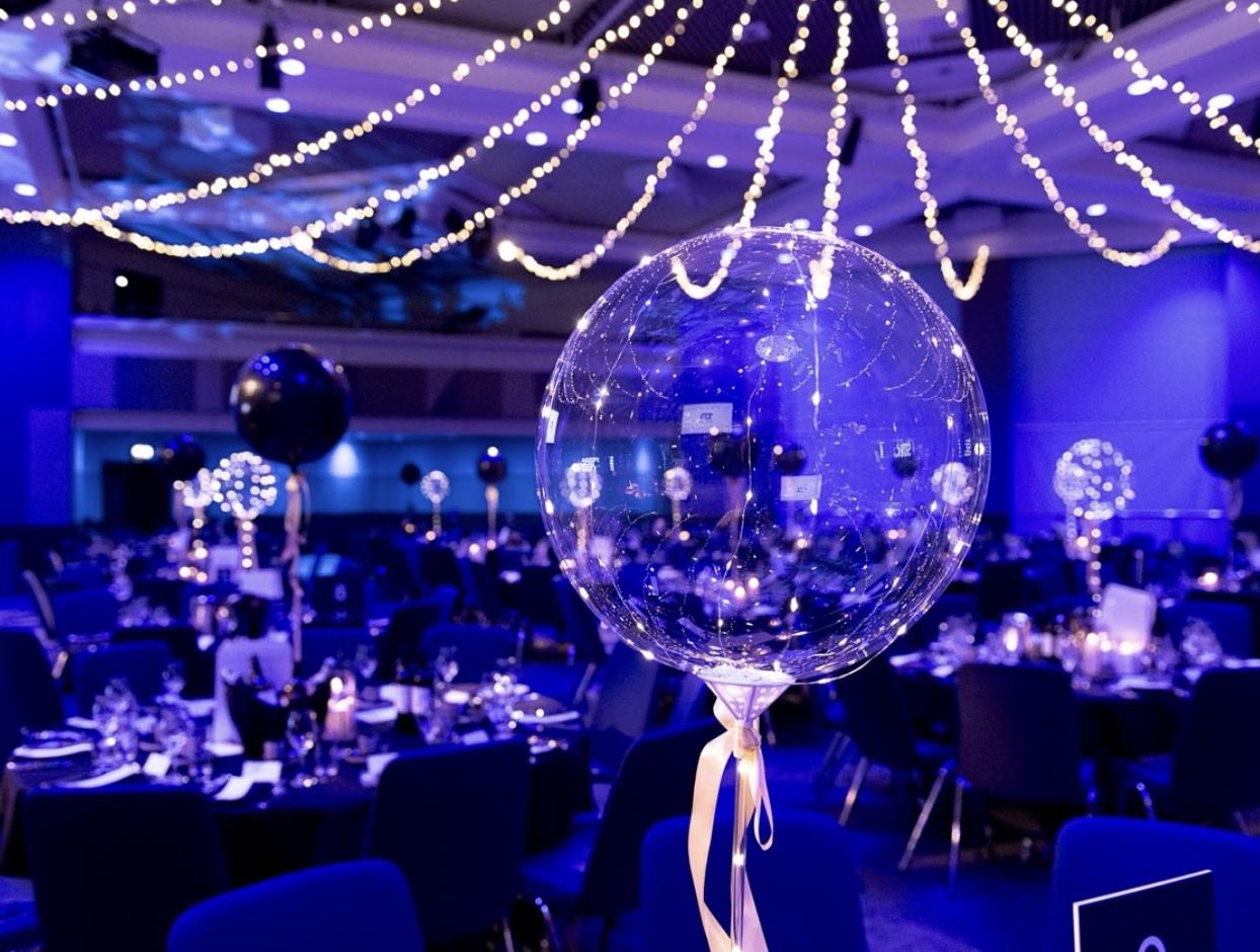 The event hall at ICC Birmingham arranged with dining tables, blue lighting, strings of lights and illuminated transparent balloon centrepieces