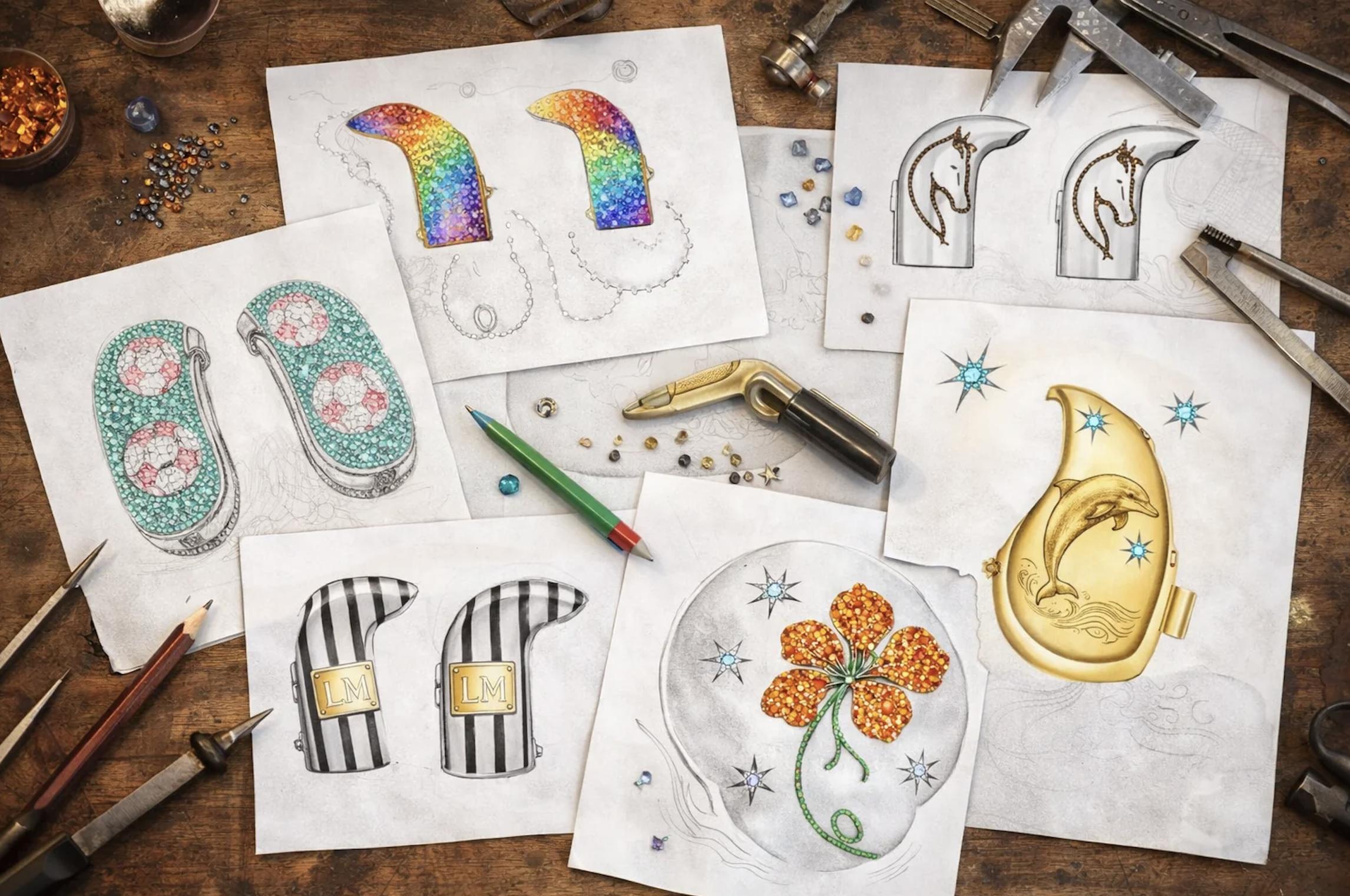 Jewellery workbench covered with AUZI hearing-device cover sketches featuring rainbow gemstones, flowers, dolphins, horses and striped designs, alongside tools and loose stones