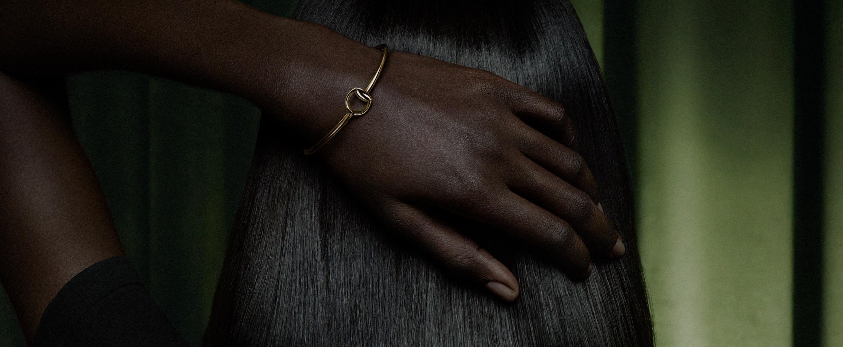 Close-up of a model wearing a slim polished yellow-gold Gucci Horsebit bangle against a dark green background
