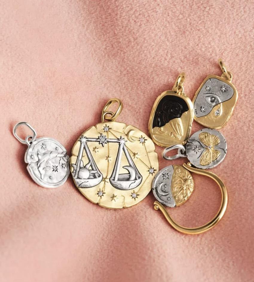 Gold and silver Pandora Talisman charms arranged on pink fabric, including a large Libra medallion, celestial symbols and a butterfly motif