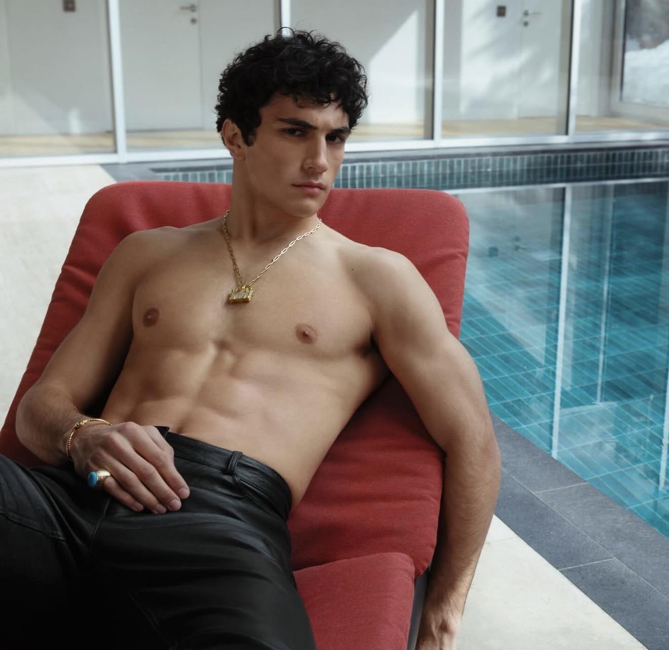 Model reclining on a red chair by a pool, wearing a gold pendant necklace, ring and bracelet. NeverNot Jewellery