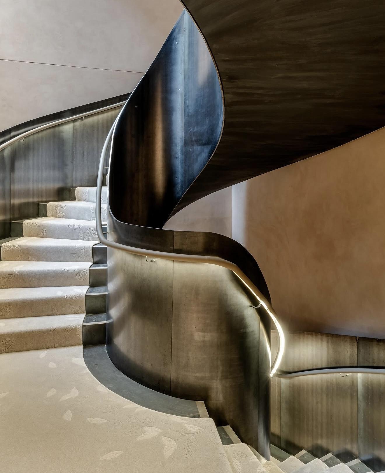 Curving staircase inside The Boodles Townhouse with dark metal balustrades, illuminated handrails and a pale carpet decorated with a subtle leaf pattern
