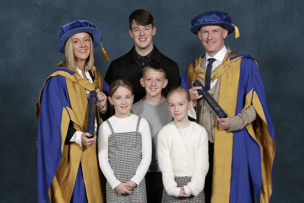 Amy and Ryan wearing blue and gold academic robes and holding certificate tubes, posing with four children against a dark blue backdrop