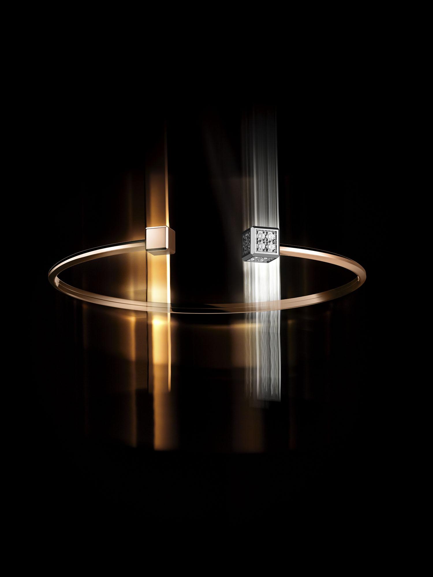 A gold bangle featuring two cube elements&mdash;one plain gold and one diamond-set&mdash;set against a dark background with light reflections highlighting their geometric form.
