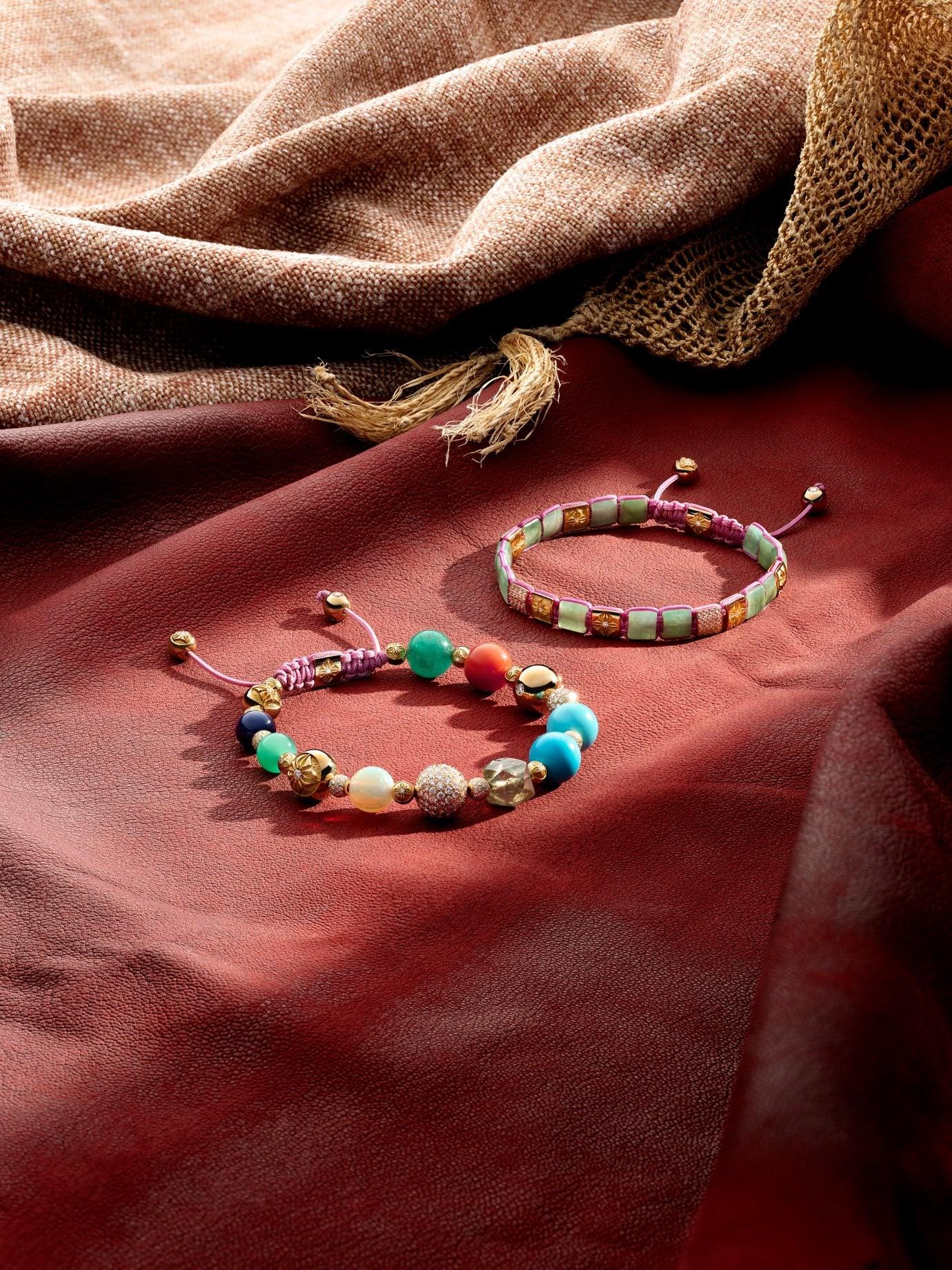 Two colourful Shamballa Jewels bracelets featuring polished gemstone beads, gold details and pink macramé braiding, arranged on deep red fabric
