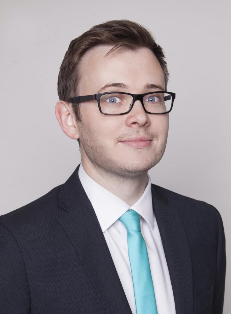 Portrait of John Watson of PJ Watson, wearing glasses, a dark suit and turquoise tie