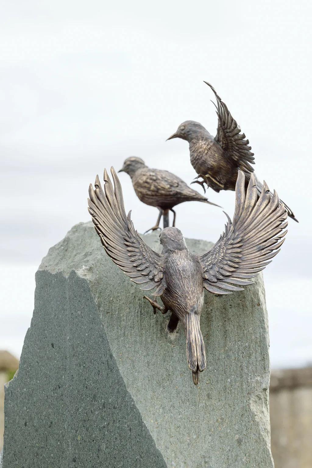 Close-up of three detailed bronze bird sculptures, two with wings spread, positioned at the top of a tall grey stone memorial