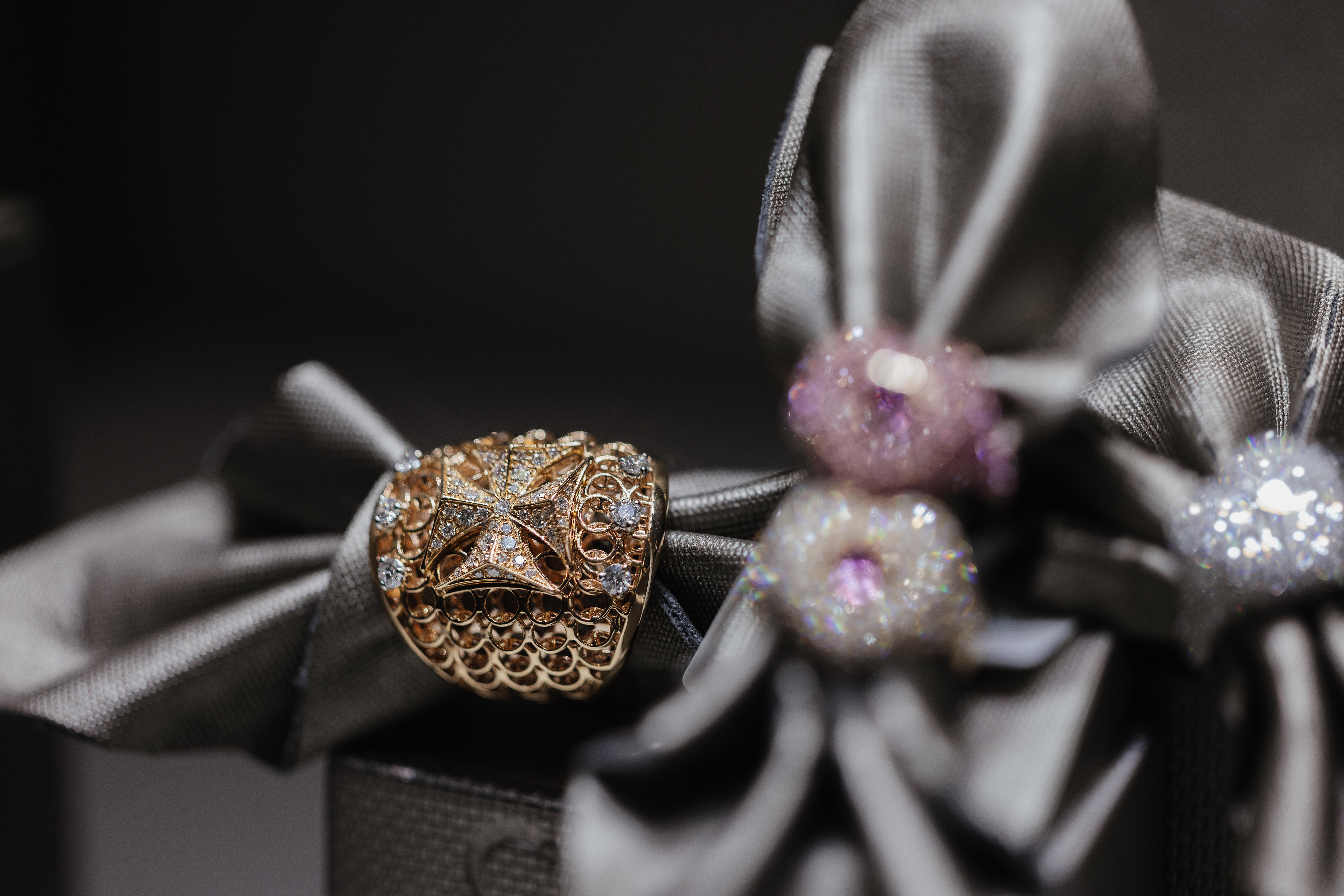 Close-up of a wide gold openwork ring set with colourless stones, displayed on dark metallic fabric beside sparkling beads.