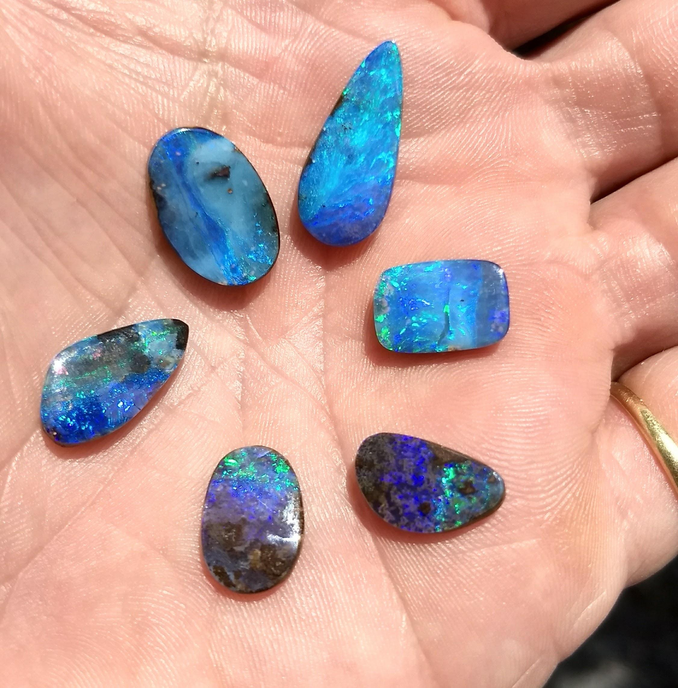 Six polished Queensland boulder opals resting in a hand, displaying freeform shapes and vivid blue, green and violet play-of-colour
