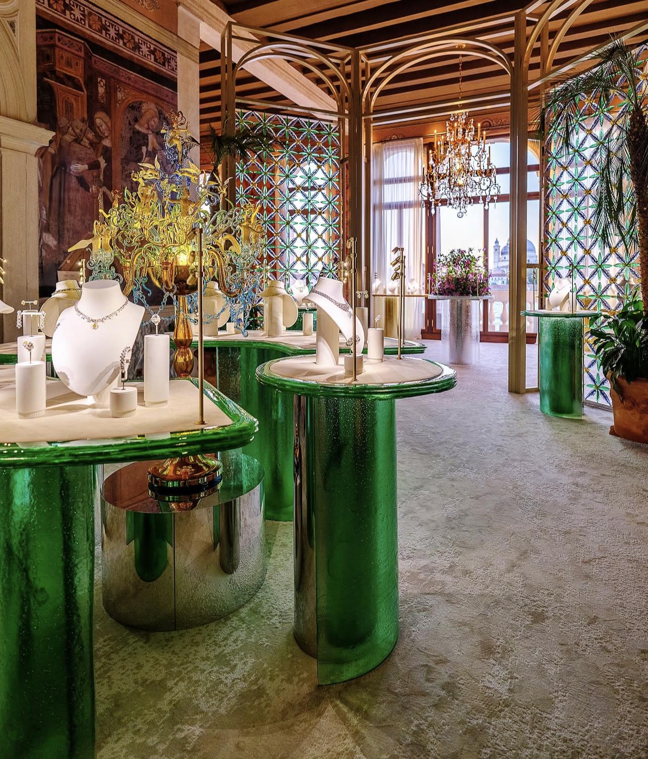 Wide view of Tiffany’s Venice exhibition with green glass tables, jewellery displays, chandeliers and botanical screens.