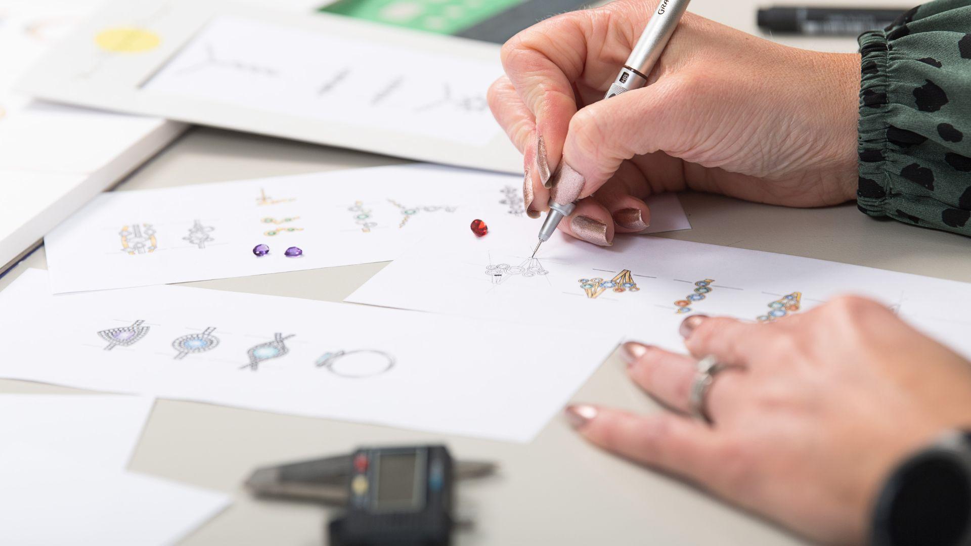 Jewellery designer sketching a coloured gemstone design beside drawings and loose gemstones.