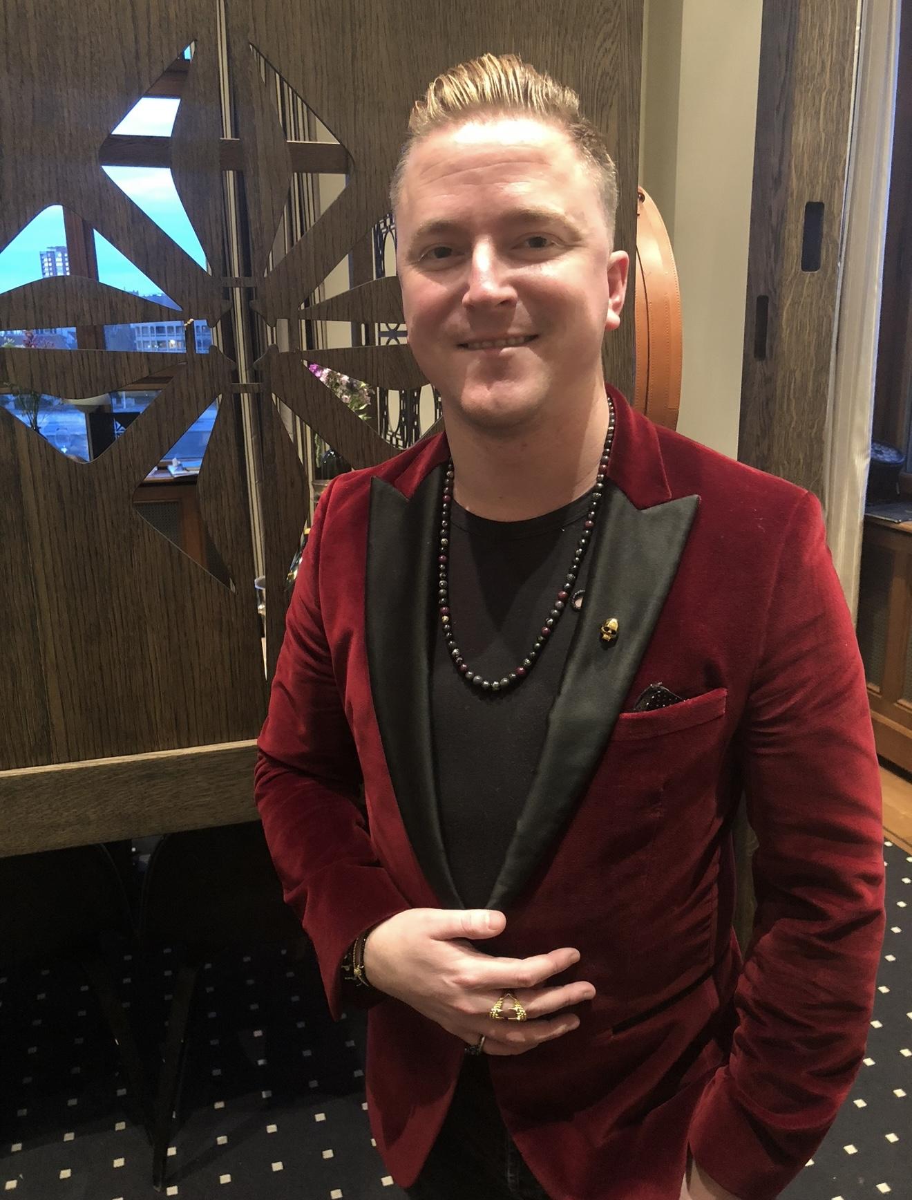 Andrew Martyniuk at Baselworld in 2018 wearing a red velvet jacket with a Shamballa Jewels beaded necklace, bracelet and rings