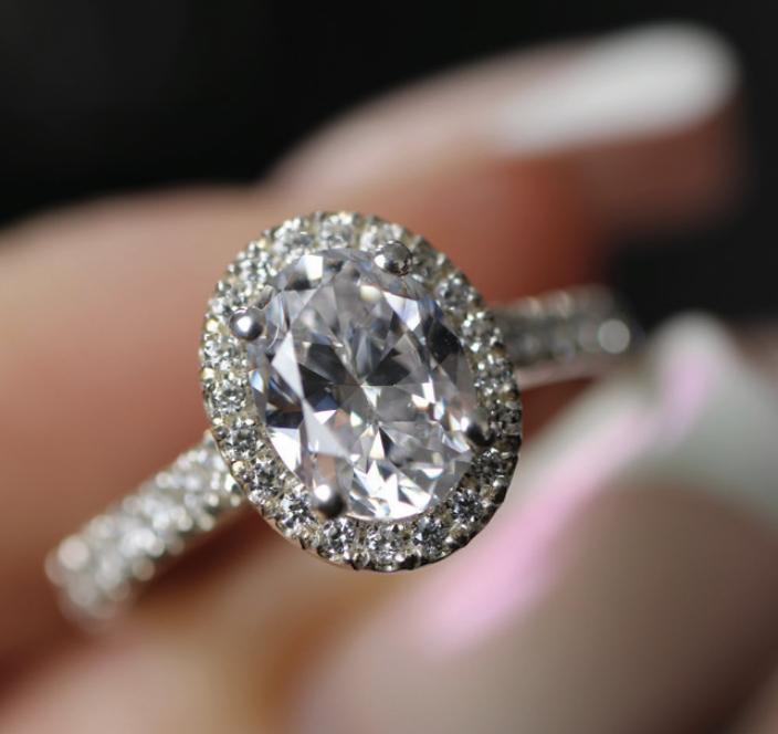 Close-up of an oval-cut diamond halo ring with a pav&eacute;-set band held between two fingers.