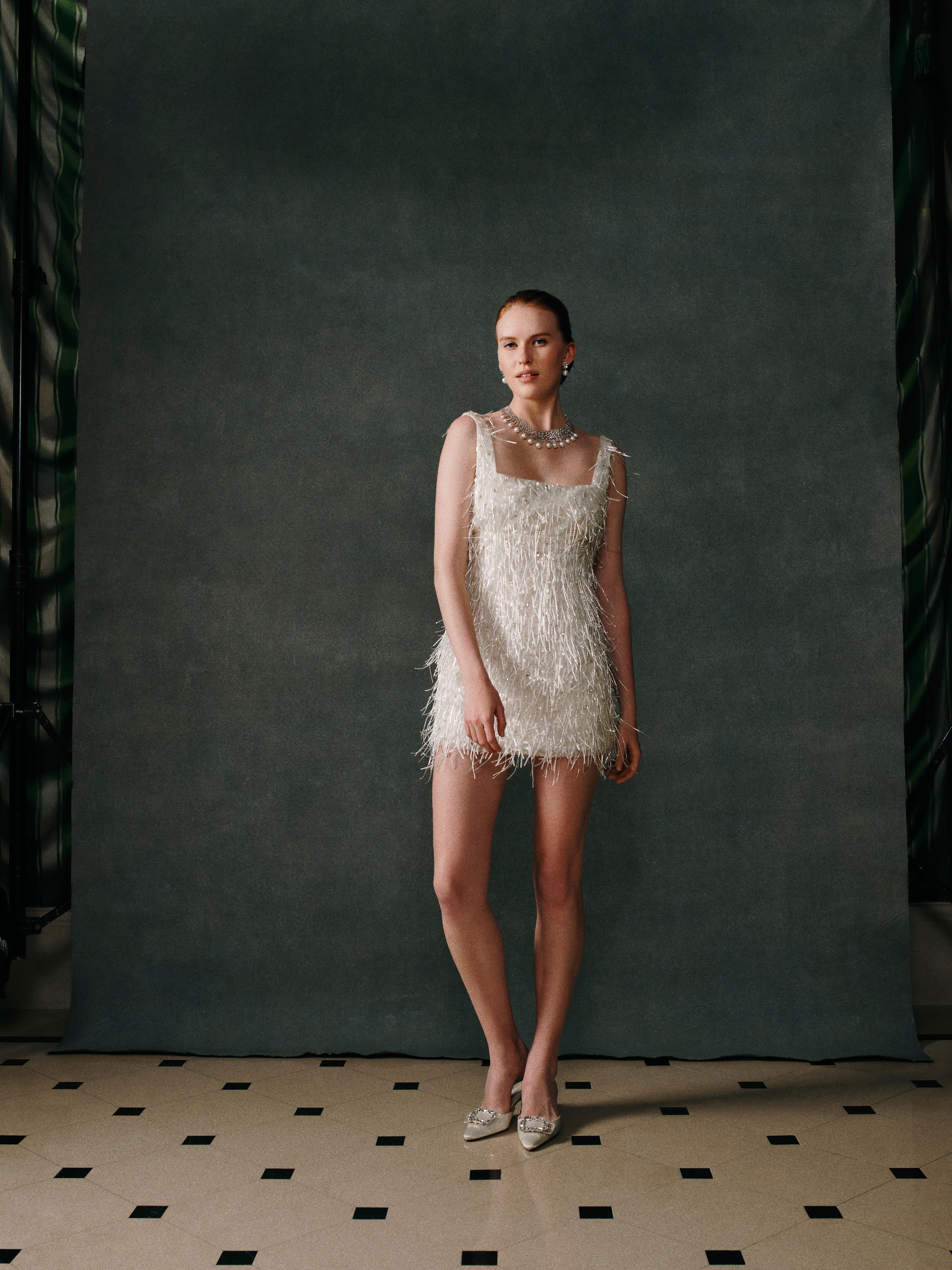 Model wearing a white embellished mini dress paired with a statement pearl necklace and matching pearl earrings from the Yoko London x Emilia Wickstead bridal collection, photographed in an elegant interior setting