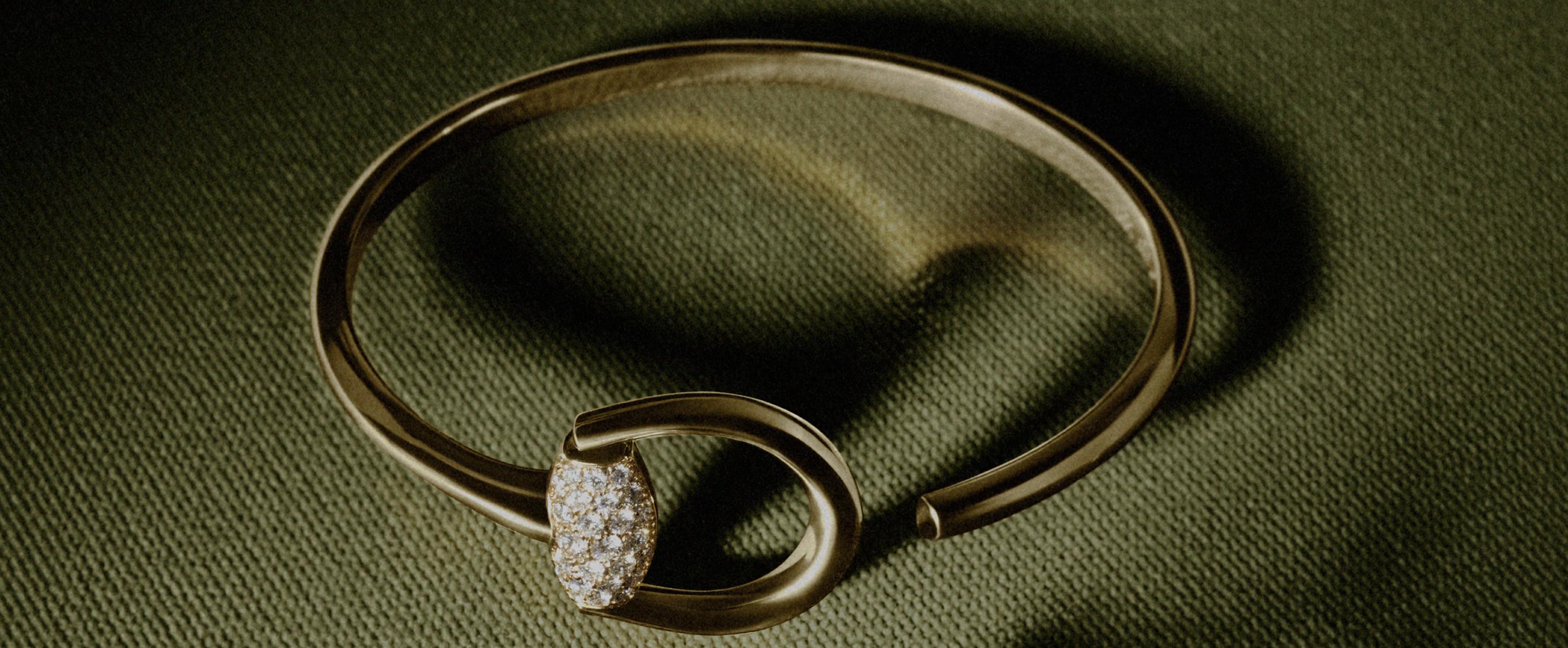Diamond-set yellow-gold Gucci Horsebit bangle arranged on textured dark green fabric.