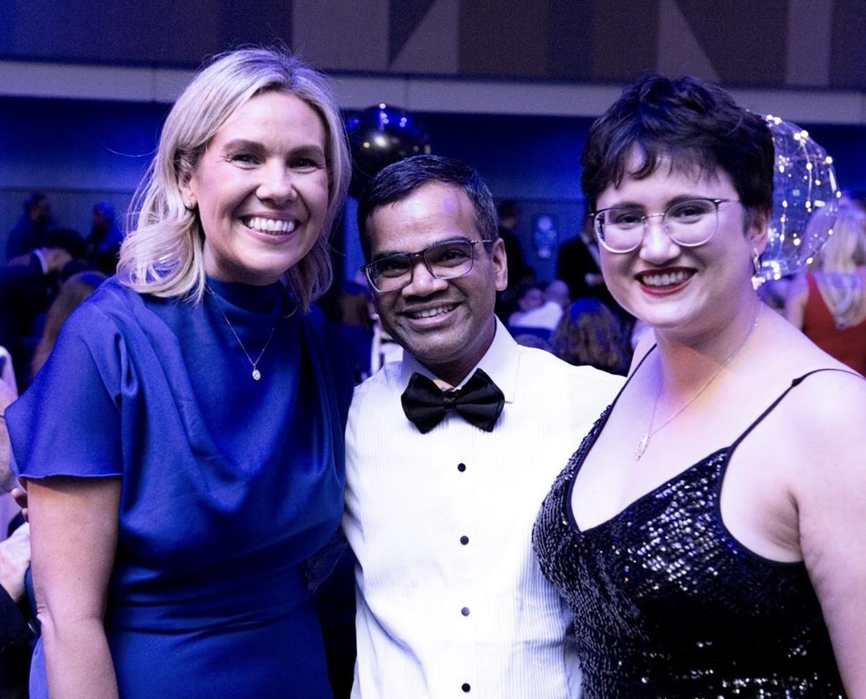 Three guests smiling together in evening dress beneath blue lighting at the NAJ Awards 2025