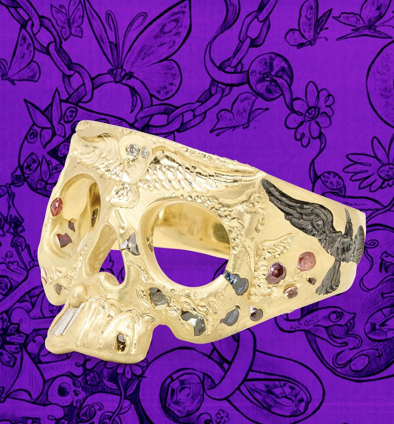 Gold skull ring from the Polly Wales x Castro Smith collaboration, decorated with engraved birds, gemstones and textured detailing against a purple illustrated background.